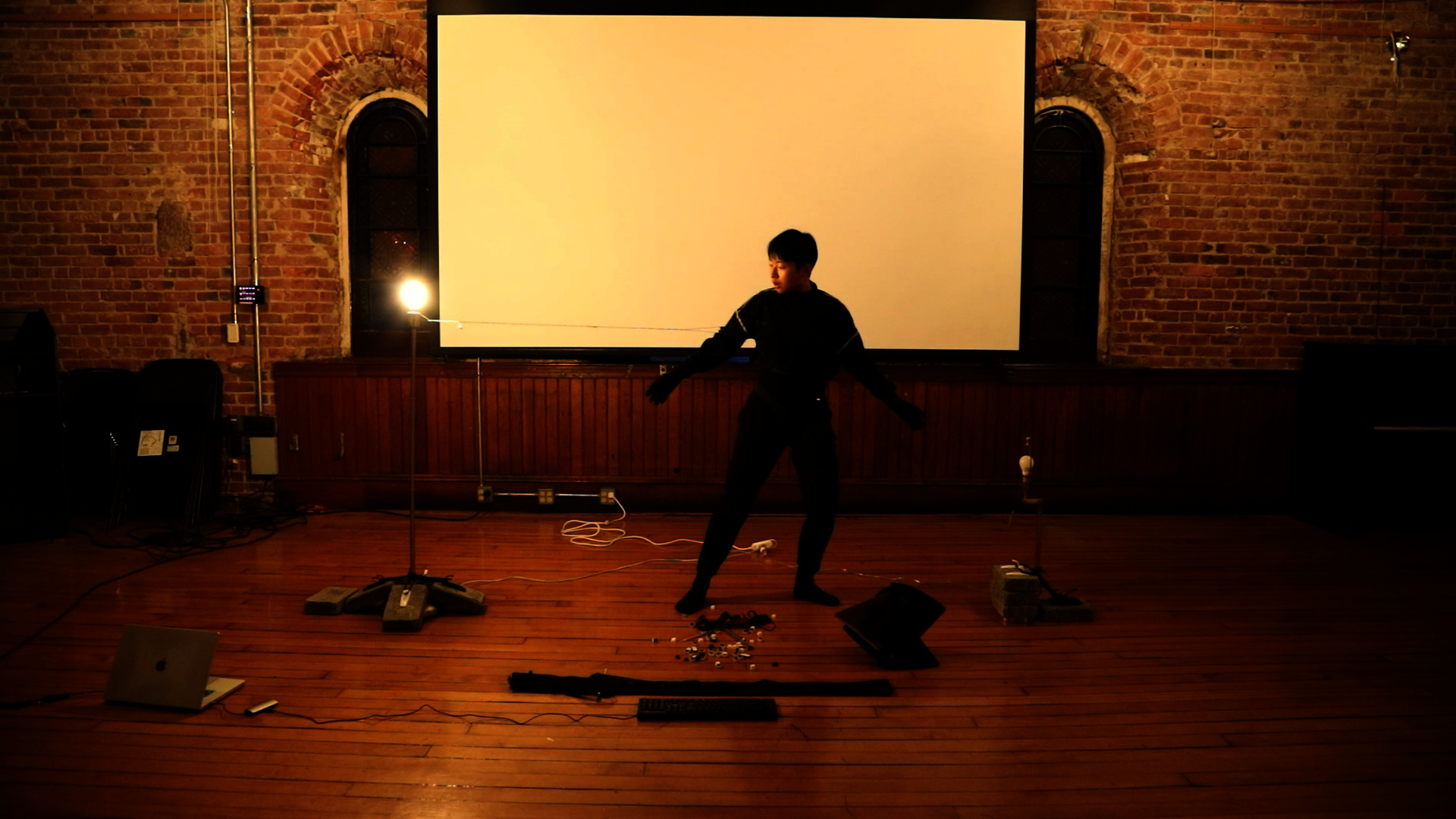 Performer in black stands among cables, props, and projection equipment in a brick-walled room.