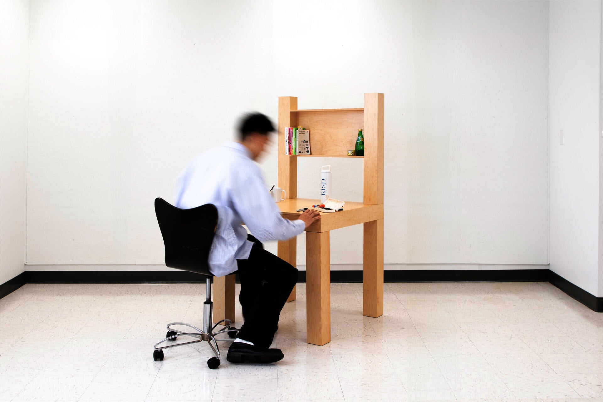A large wooden chair-shaped object with a desk surface, drawer, and shelving, functioning as a desk instead of a chair.
