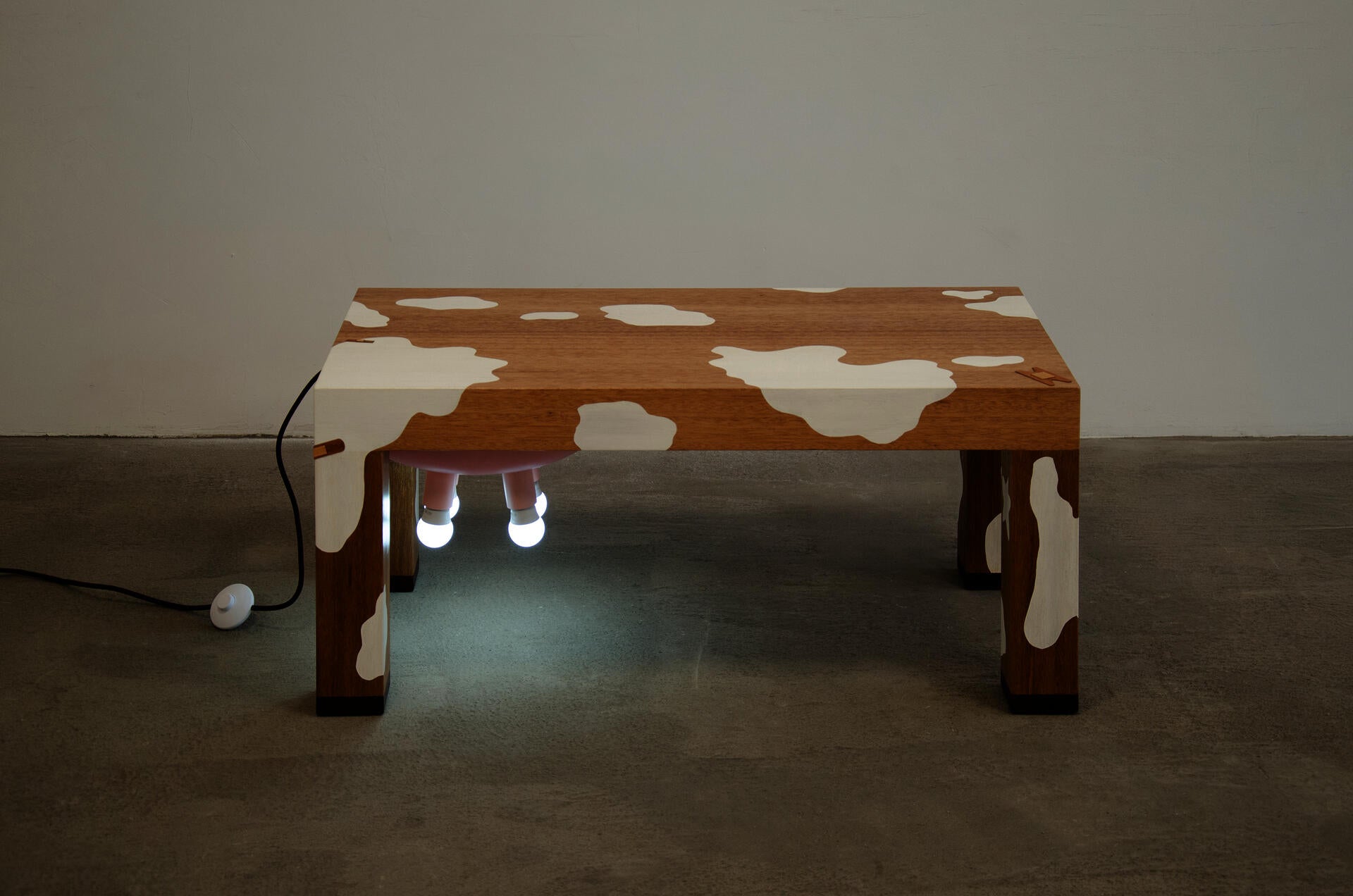 A cow-inspired wooden coffee table with a brown body, a pale pink belly, four pink light bulbs underneath like udders, and a cord extending from the back like a tail.