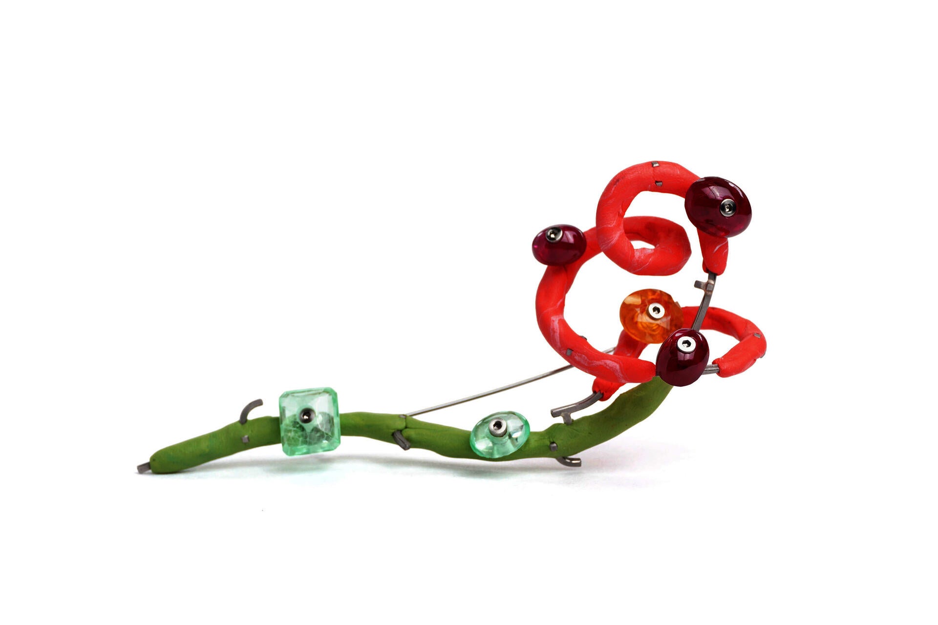 A brooch loosely in the shape of a rose, made up of marbled pink and green silicone, and metal wire. Rubies and sapphires are bolted along its length with screws. On a white background.