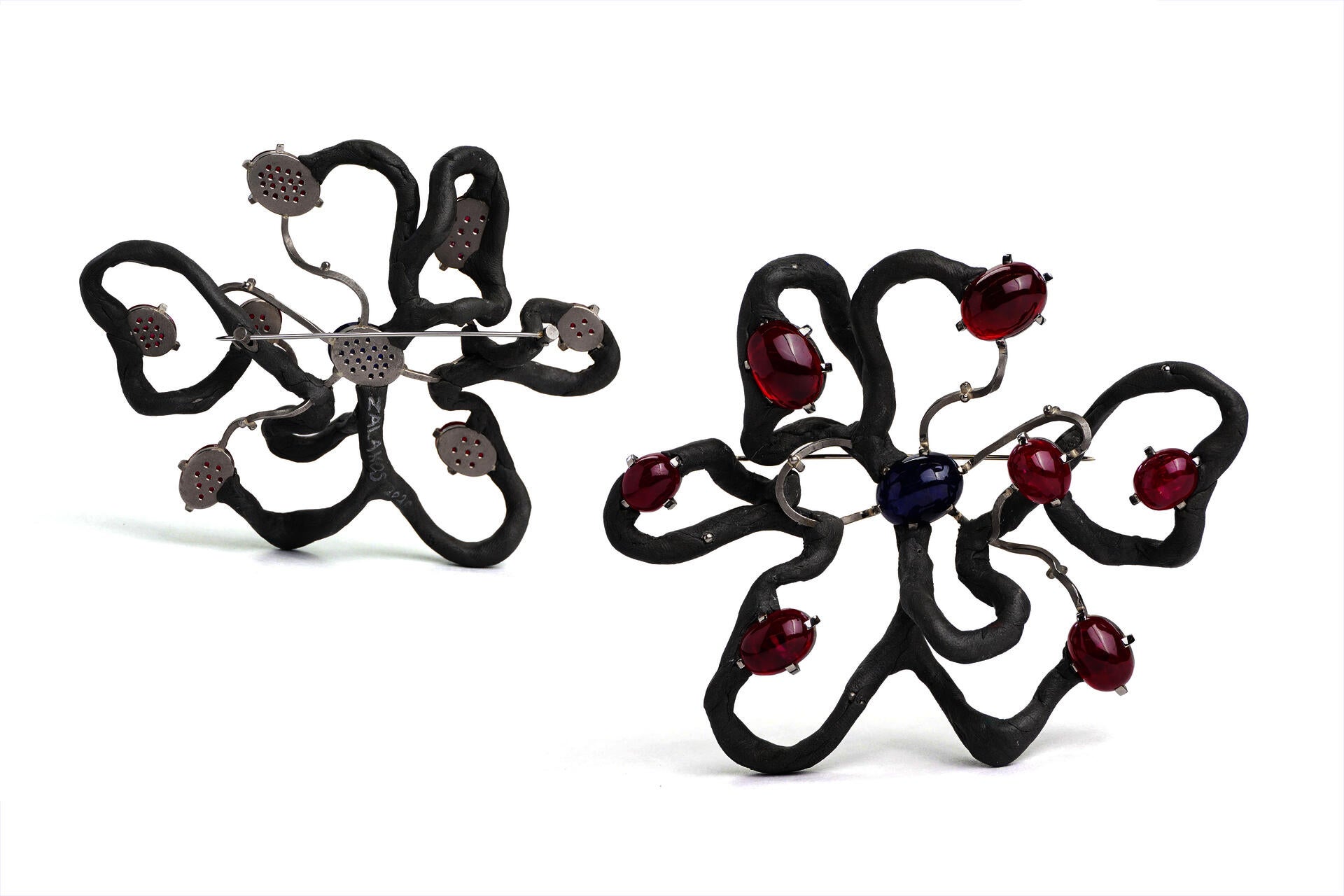 A brooch loosely in the shape of a flower with seven large petals, made up of grey epoxy and metal wire. Rubies and sapphires are set in prong settings along its length. The brooch is pictured