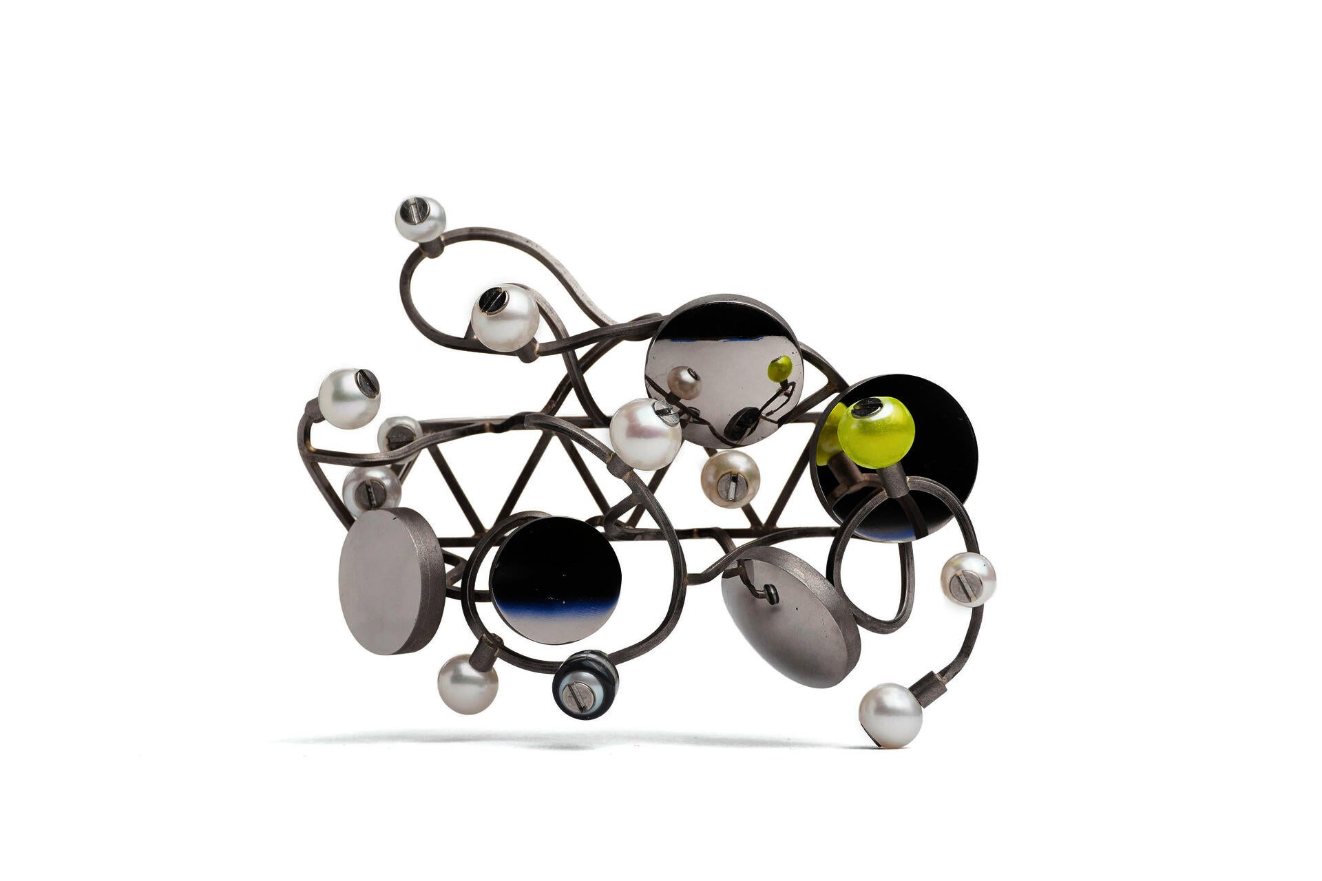 A brooch loosely in the shape of a triangular truss with branching and twisted lengths of wire wrapped around it. Circular mirror panels and pearls are bolted along its length with screws. On a white background.
