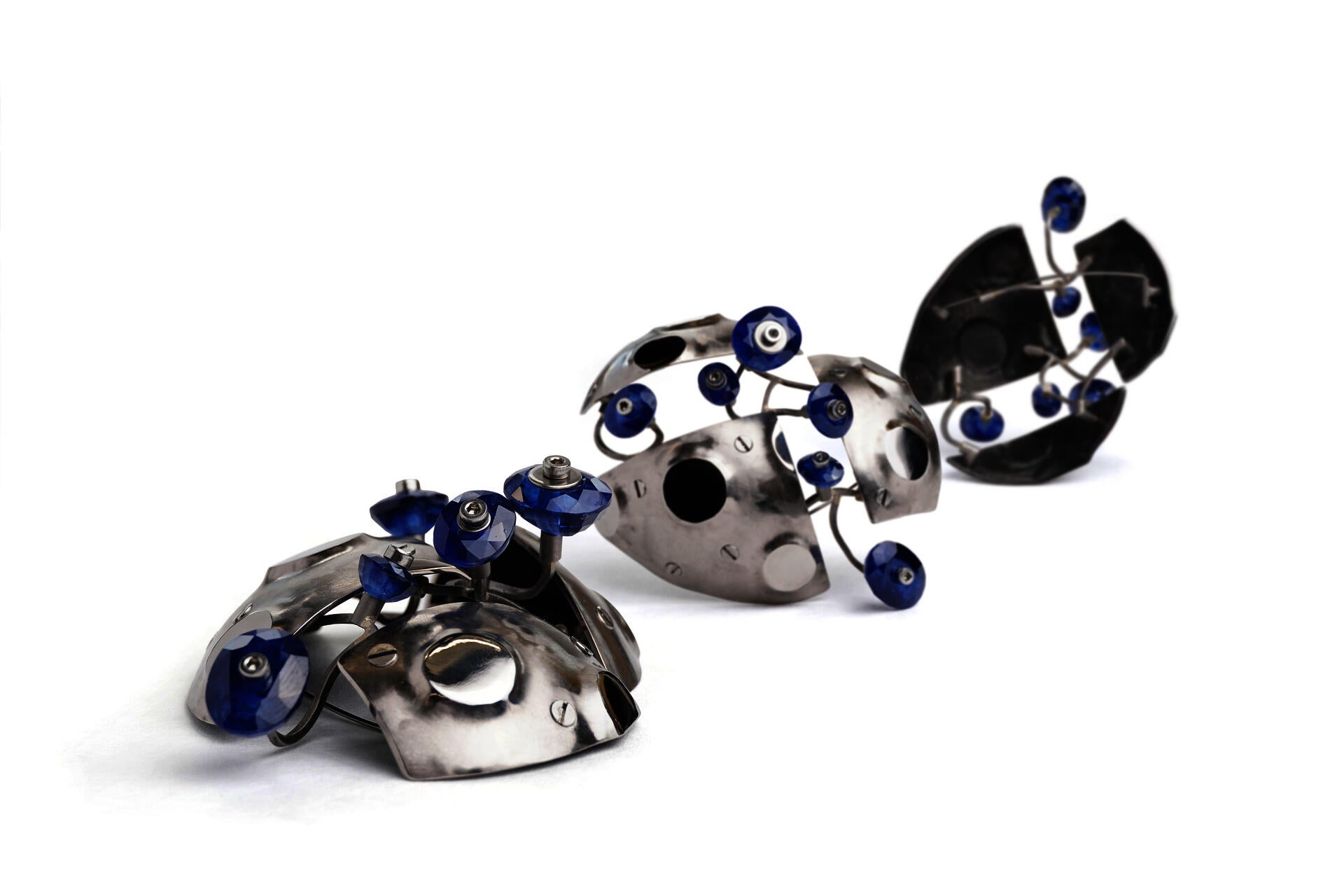 A brooch made from three rounded grey metal panels that are bolted together with gaps between them. Small circular mirror panels are raised from its surface with polished faces. Short lengths of twisted square wire branch