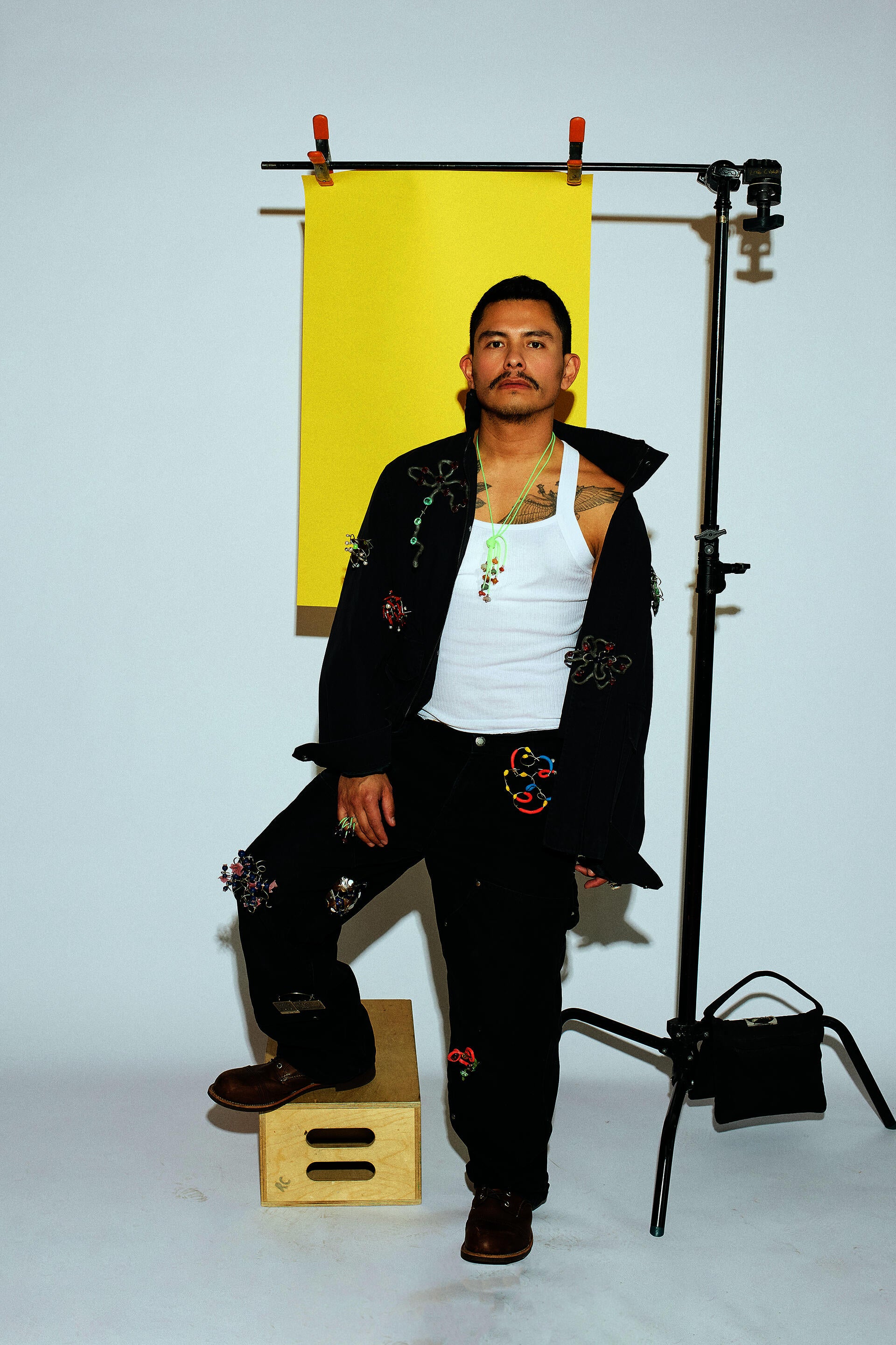 A man in workwear standing in a photo studio on a white background. A tripod beside him holds a sheet of yellow paper behind his head. He has twelve brooches pinned to his clothes and wears rings and pendants.