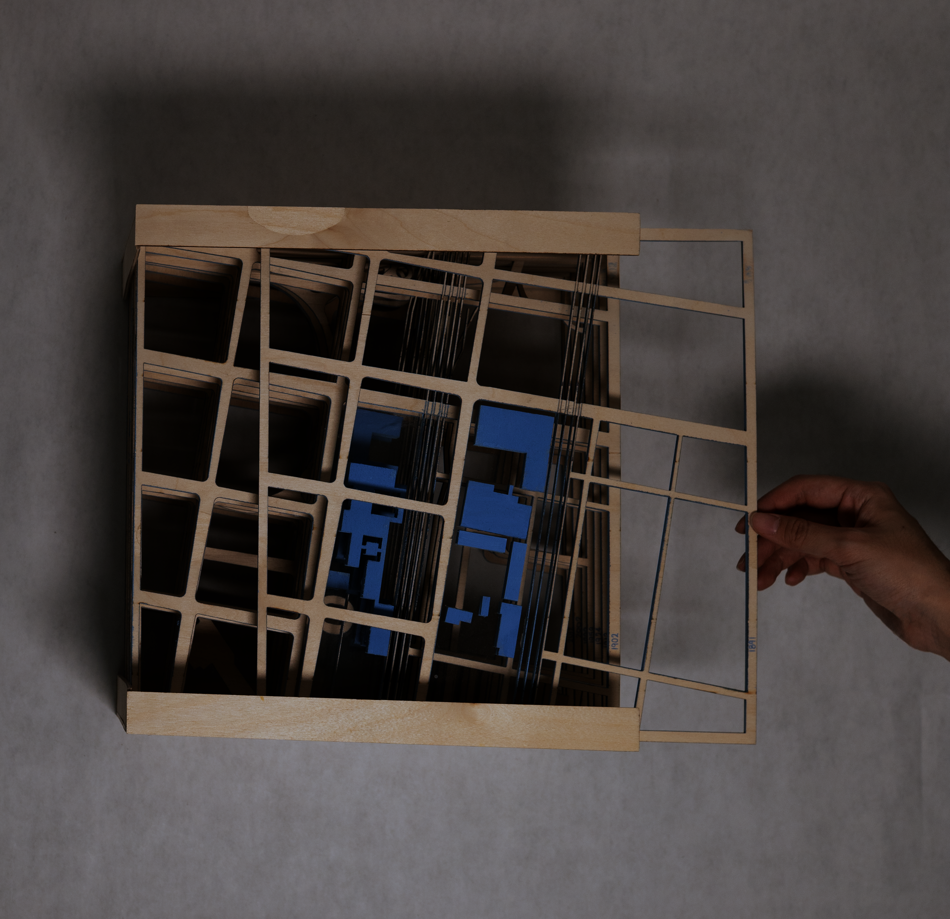 Hand pulling square wooden map with blue buildings to reveal maps of later years layered beneath.