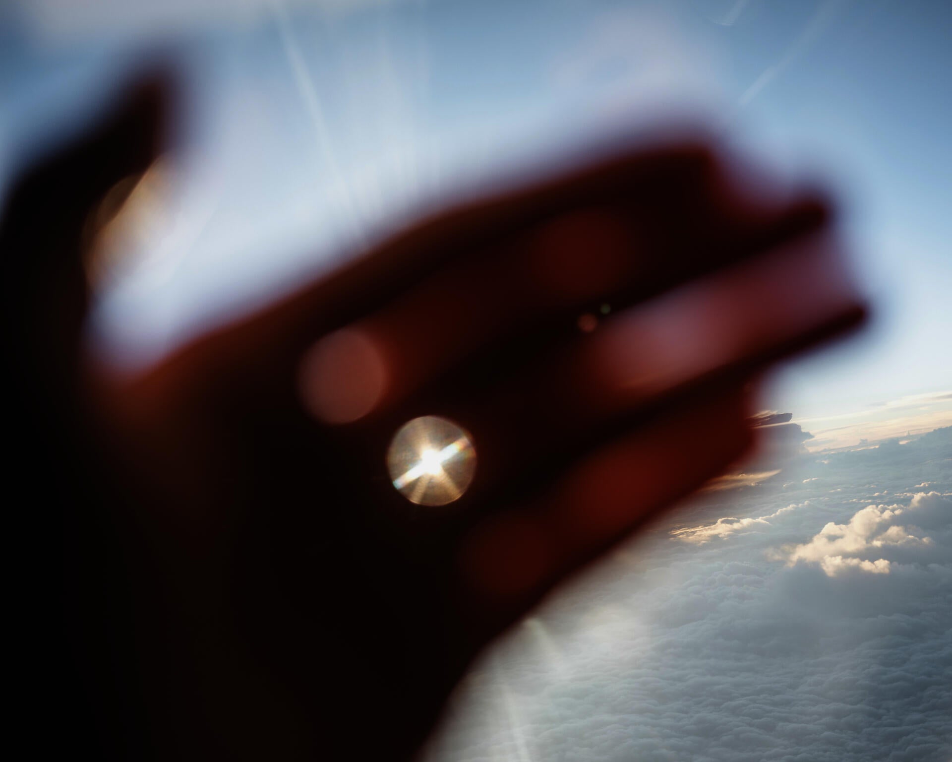 Color photograph of the palm of a hand glowing in orange, held in front of a bright blue sky, with the sun peeking through the gap between the fingers.