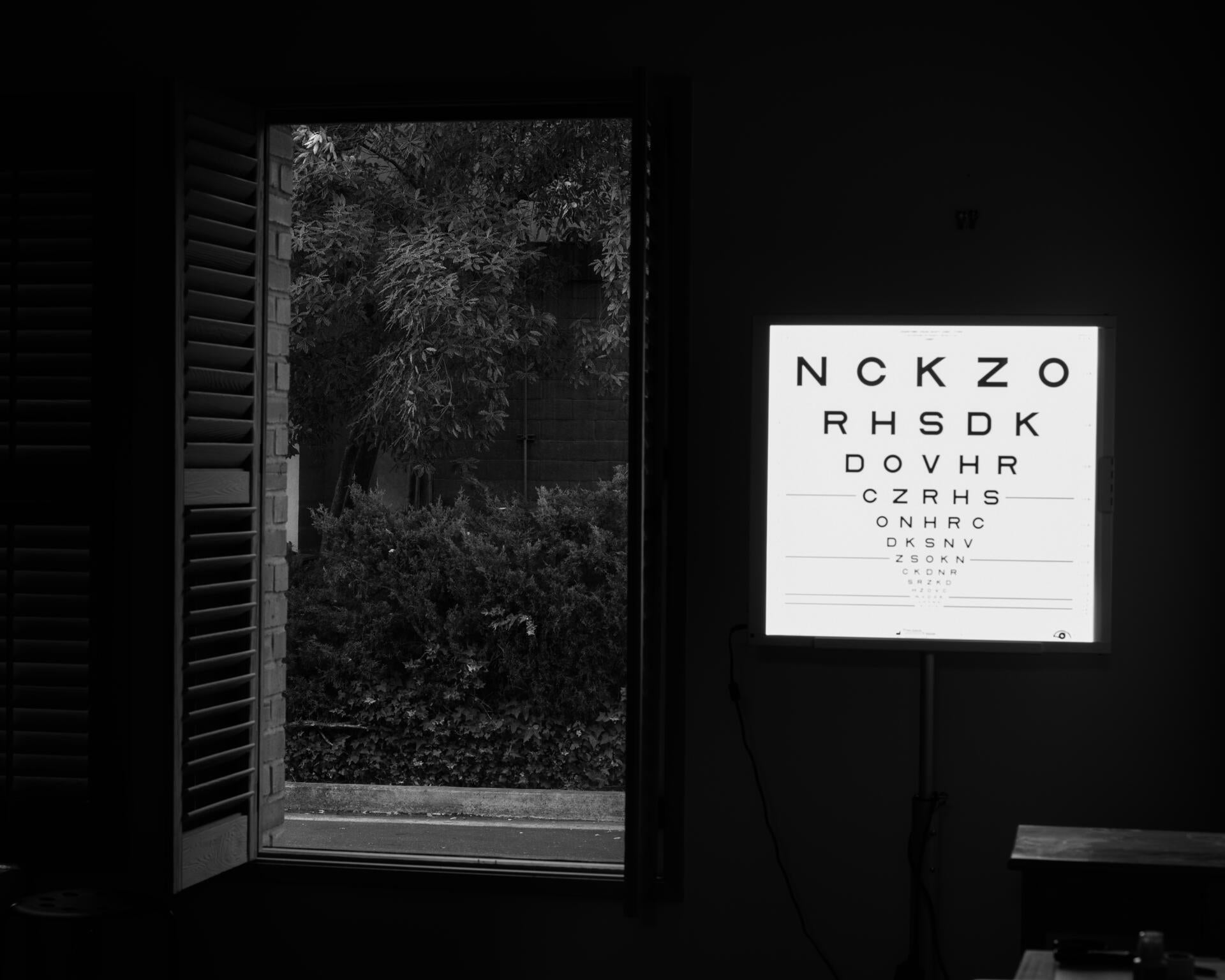 Black and white photograph of inside the vision test room. A soft glowing vision test chart illuminates the right side of the room. To the left, a tall window with its wooden blinds open, shows trees and plants outside.