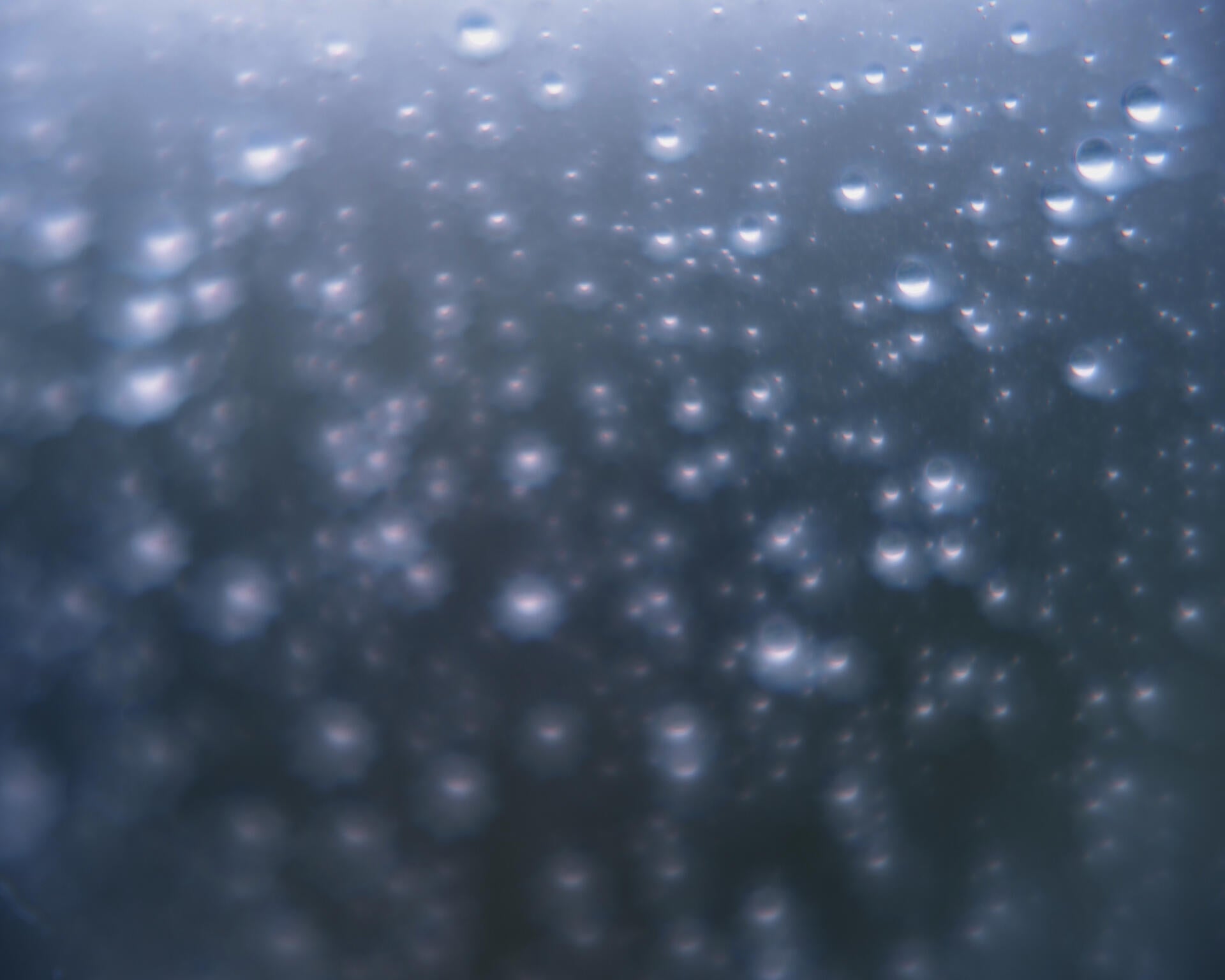 Color photograph of blurry raindrops, suspended in watery blue background.