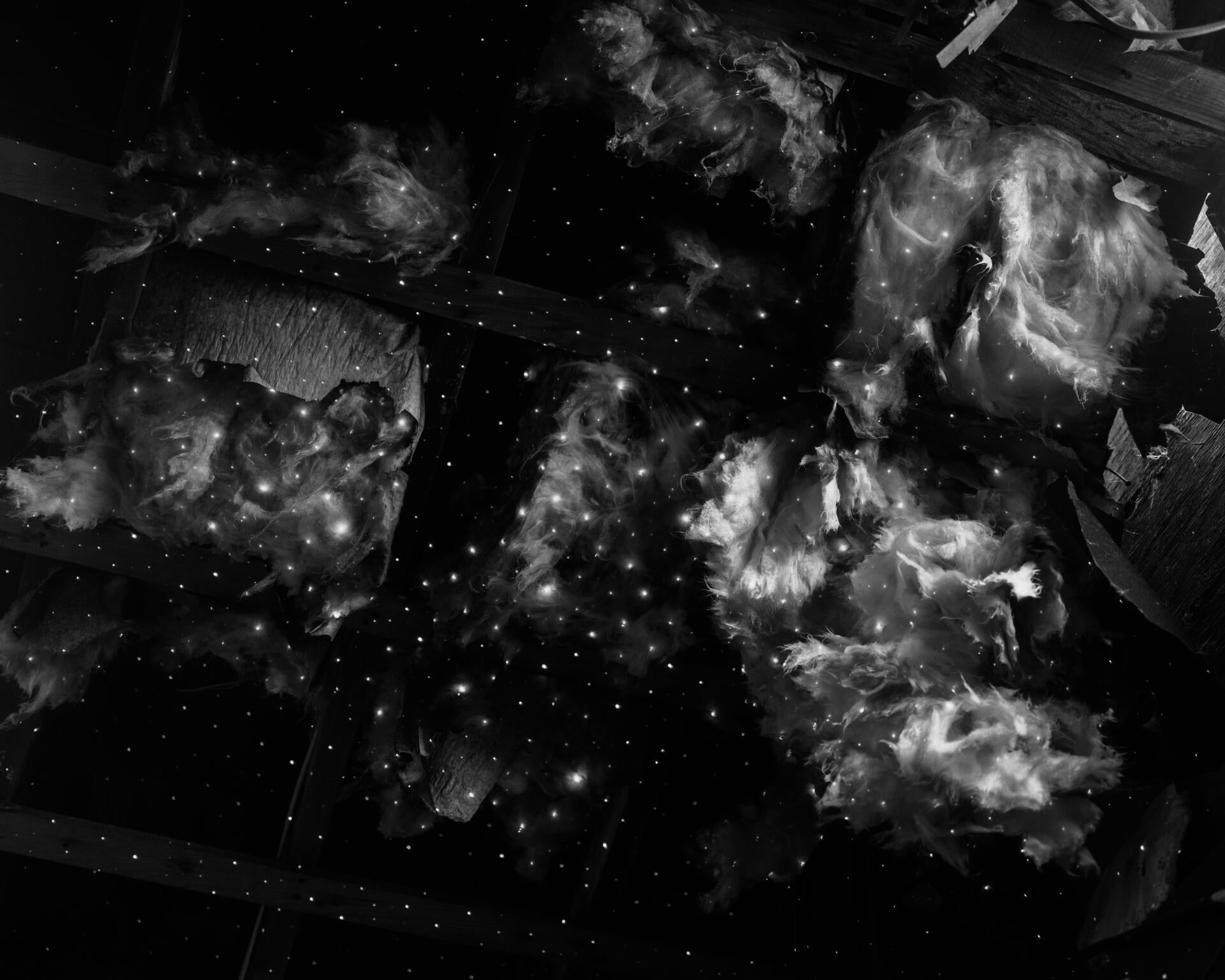 Black and white photograph of tiny white dots of light glimmering like stars, on a collapsed ceiling. Fiberglass insulation materials are falling out from the wooden frames of the ceiling.