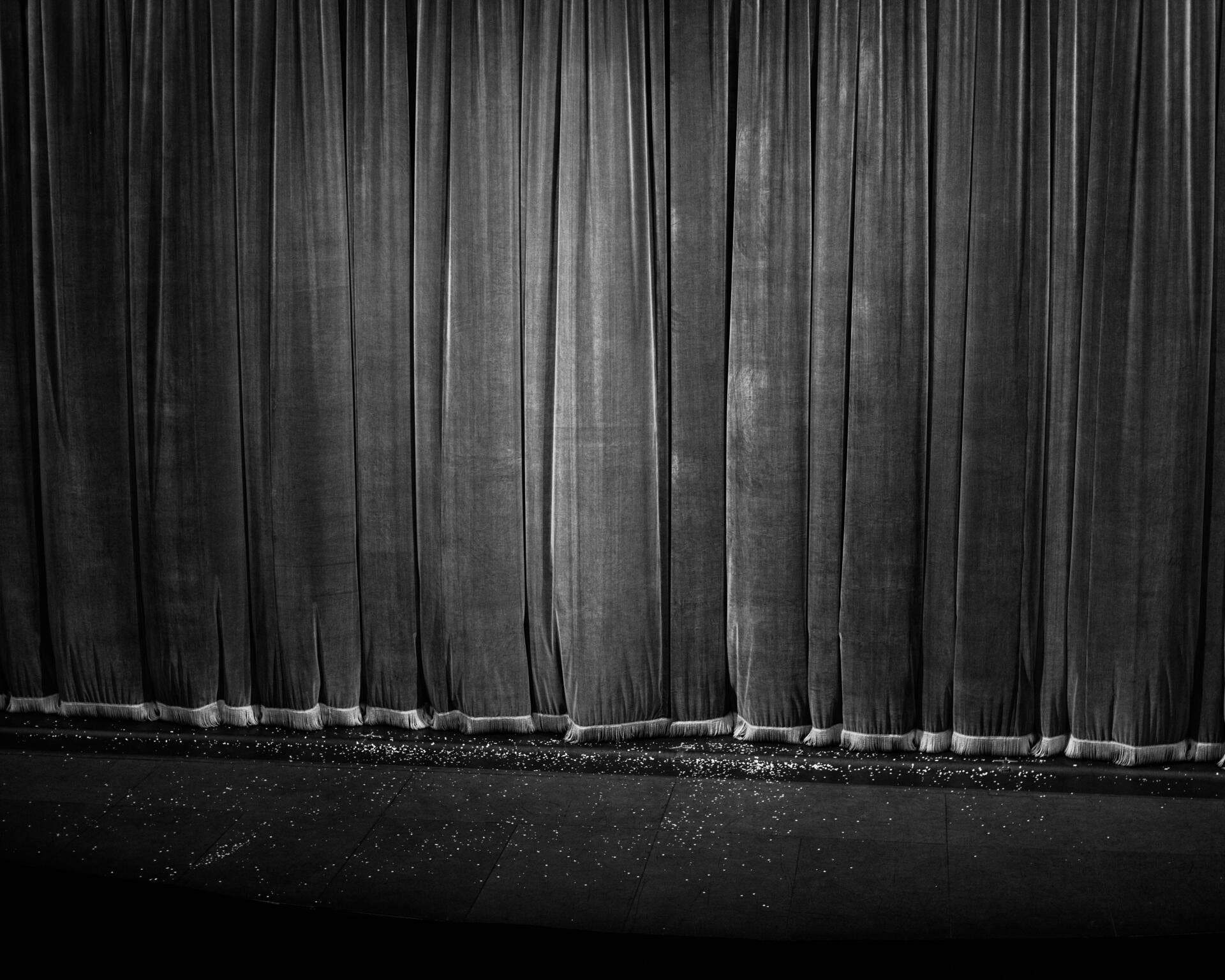Black and white photograph of a thick velvet curtain, closed, with tassels dragging on the stage floor. Small white flakes are sprinkled on the floor.