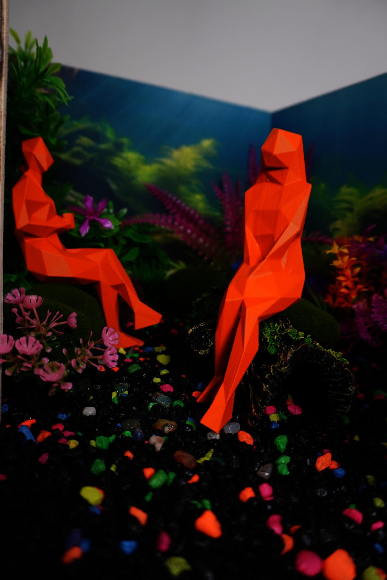Photo of a model of a waiting room designed to look like an aquarium: black gravel with neon, multi-colored pebbles mixed in; "moss" covered "rocks" and a fake, "moss" covered "log" act as seating for orange painted, angular human figures. Colorful plastic plants line the perimeter of the room and the walls are covered in a wallpaper mural of an underwater image with seaweed and sun streaks.