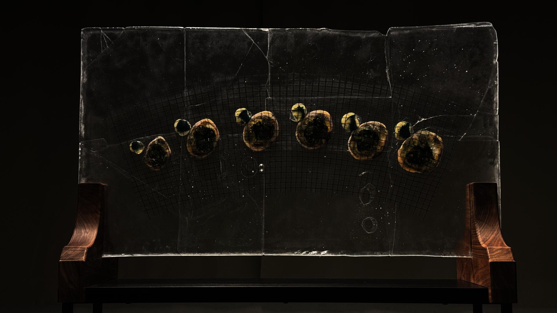 Transparent glass sheet with embedded curved glass grid and melted glass rocks held on a wooden structure resembling a distorted star map.