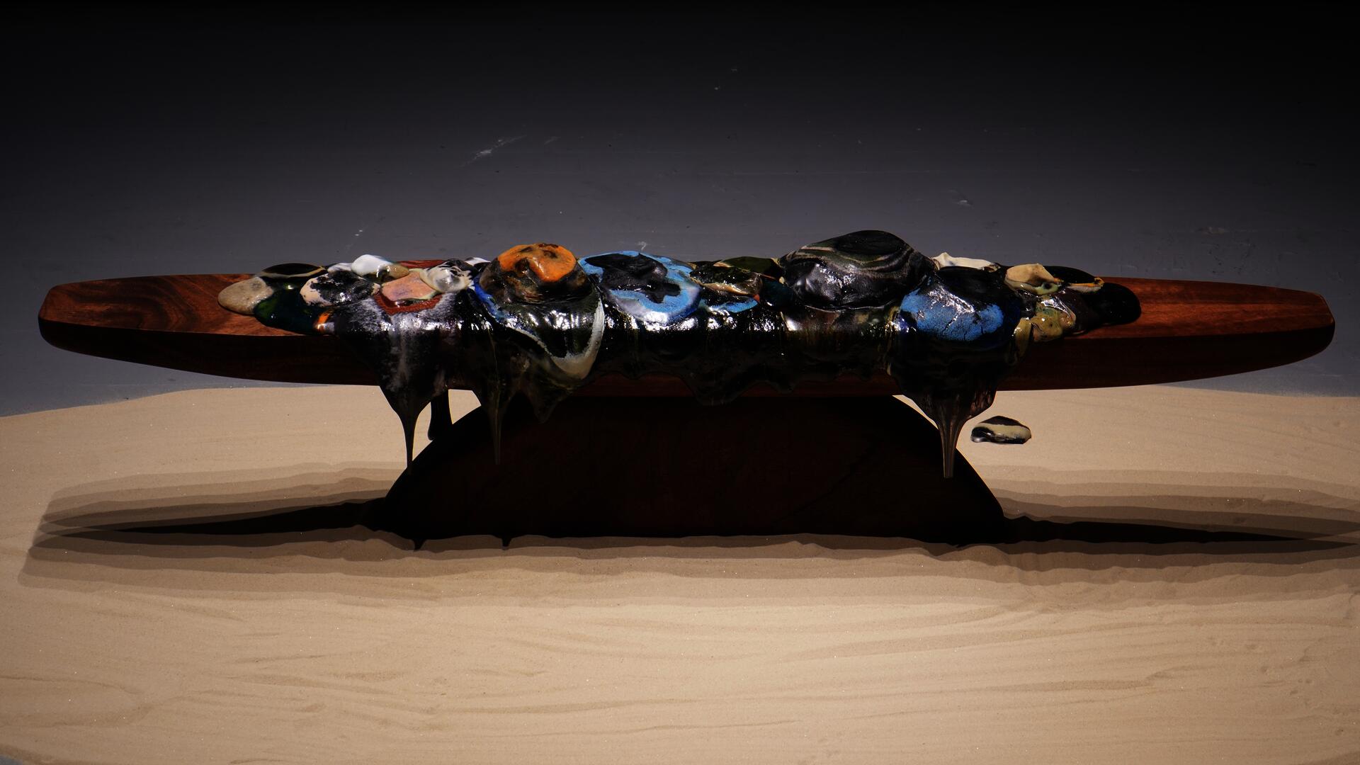 Molten glass forms in varied colors spread and drip across a long wooden platform, with softened edges and flowing material extending downward.