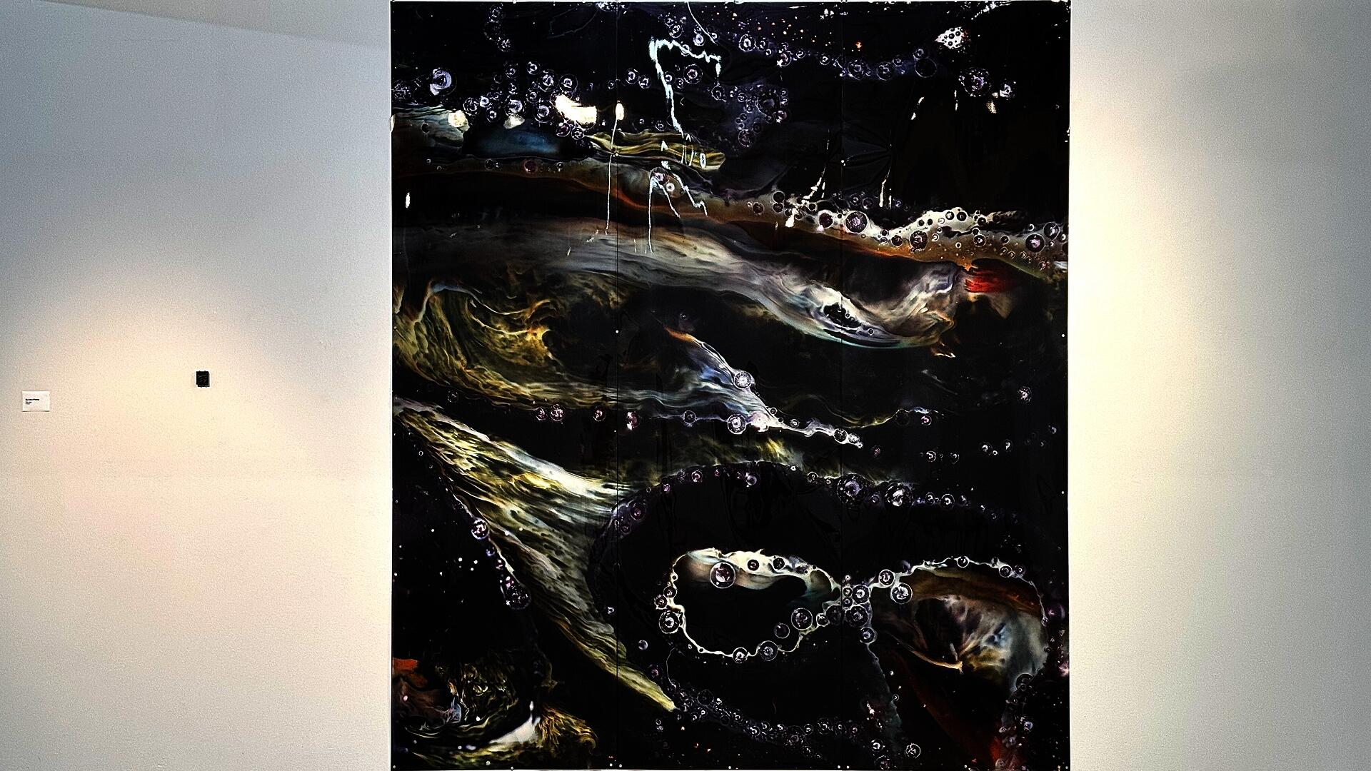 Large vertical print showing abstract patterns in dark tones with lighter flowing forms and bubble-like details, installed on a gallery wall along side with a small glass sample.