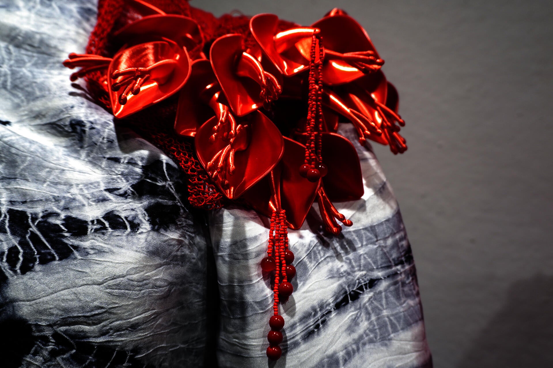 Close-up of glossy red 3D-printed ceibo flowers with beaded strands over black-and-white hand-dyed silk textile.