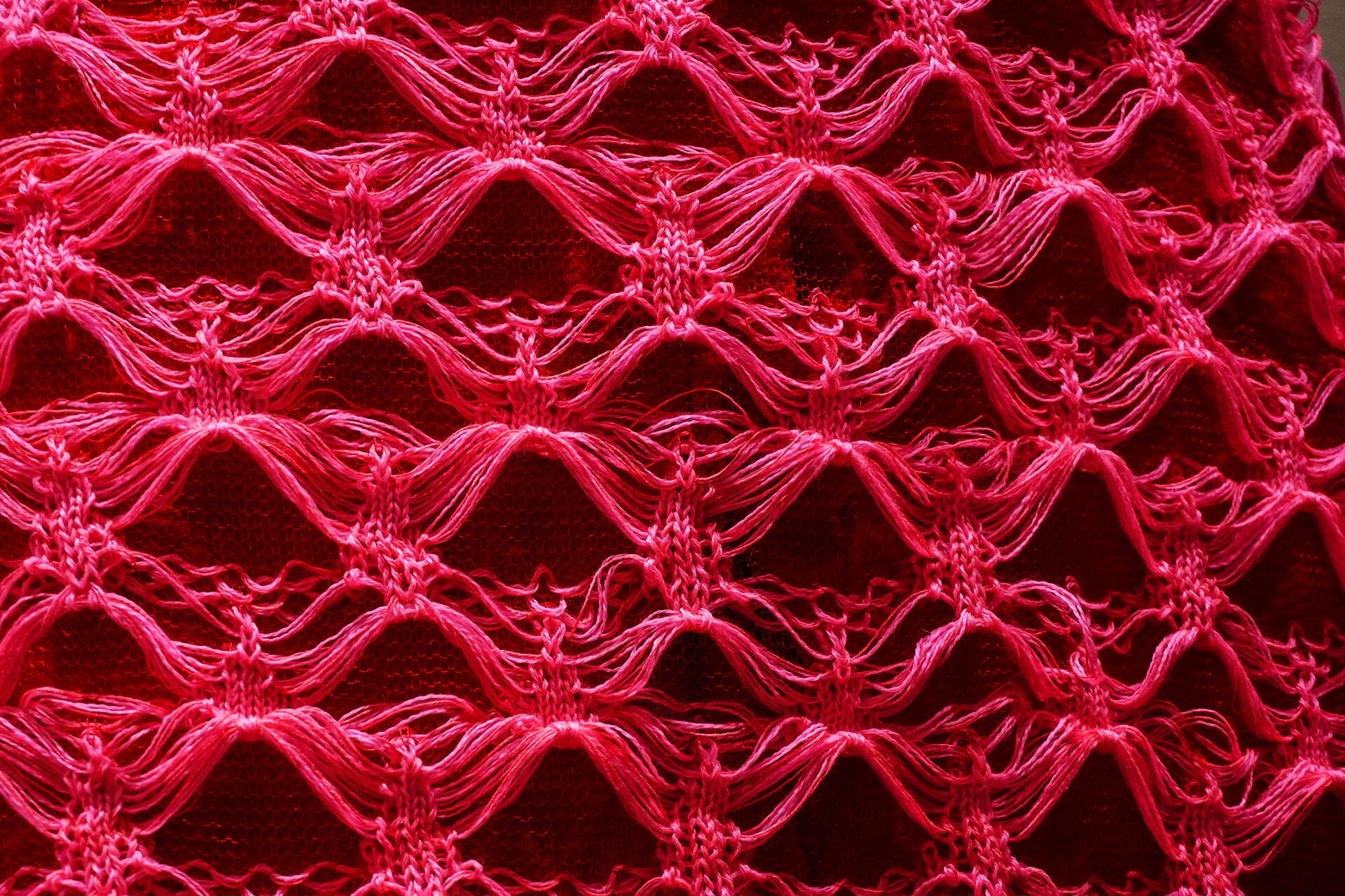 Close-up of bright pink knitted lace structure with looping openwork pattern over red textile background