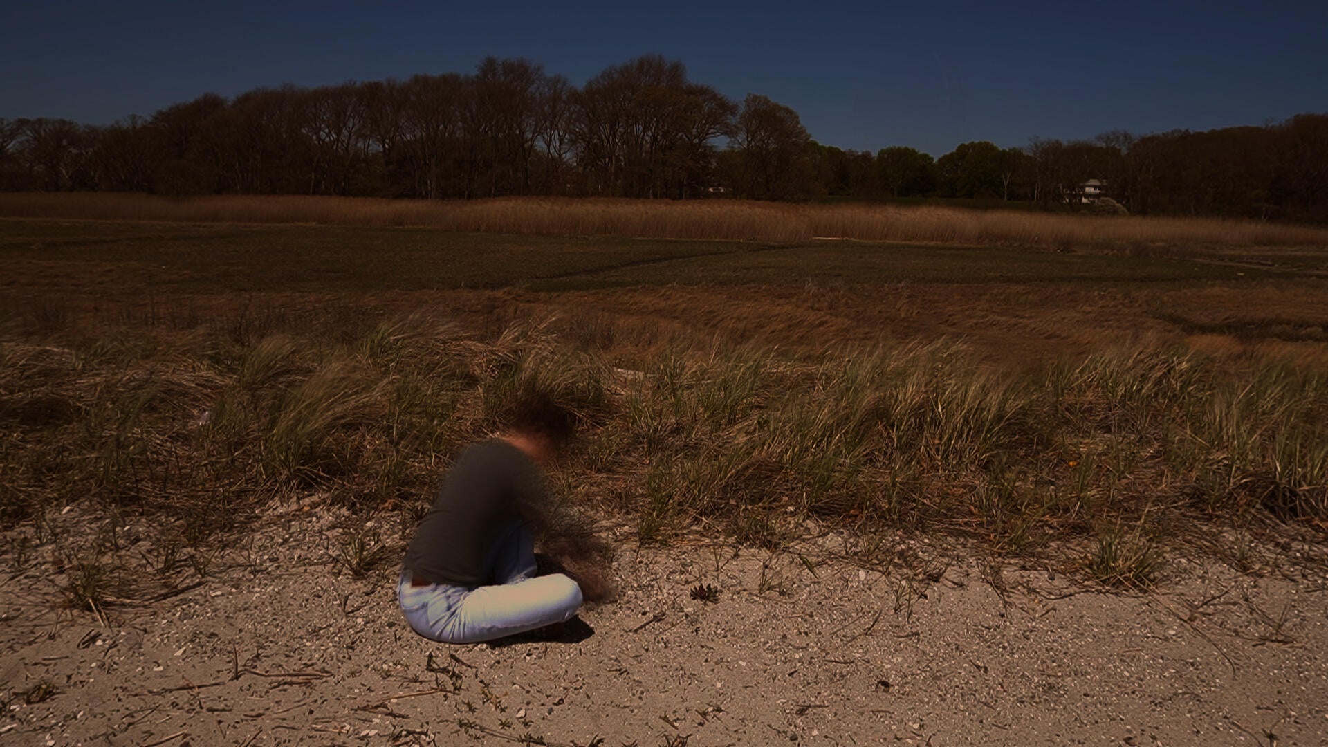 A person sits in a sandy dune landscape while making a bowl-shaped form in the ground. Marsh grasses and trees extend across the background beneath a clear sky.