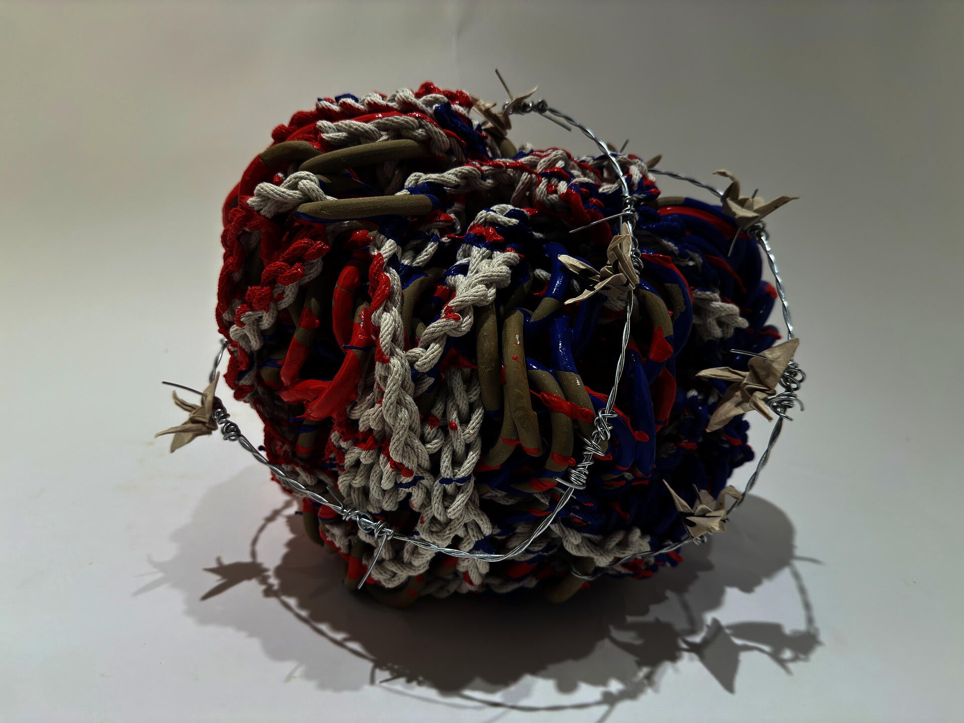 Loops smooth tan chainmail are constricted by white rope.  The piece is bathed in red and blue paint and paper cranes are spears by wrappings of barbed wire around the piece.