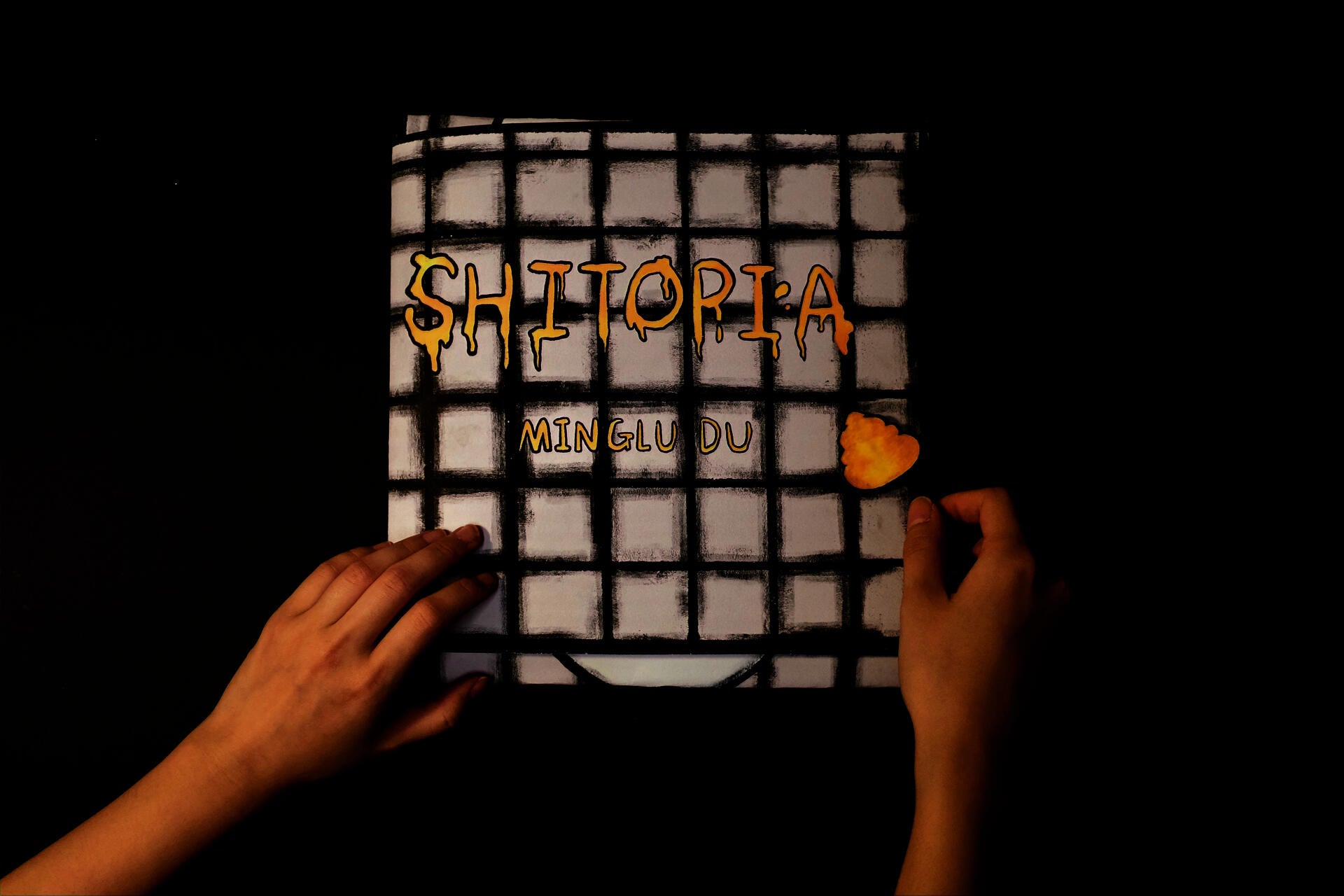 The cover of the comic book Shitopia is designed as a foldable outer shell shaped like a tiled toilet and toilet seat that wraps around the book’s contents. The cover includes the title of the work, the author’s name, and a pop-up paper clasp shaped like a piece of excrement that can be tied with string to keep the book closed. The handmade structure combines absurd humor and tactile sculptural elements.