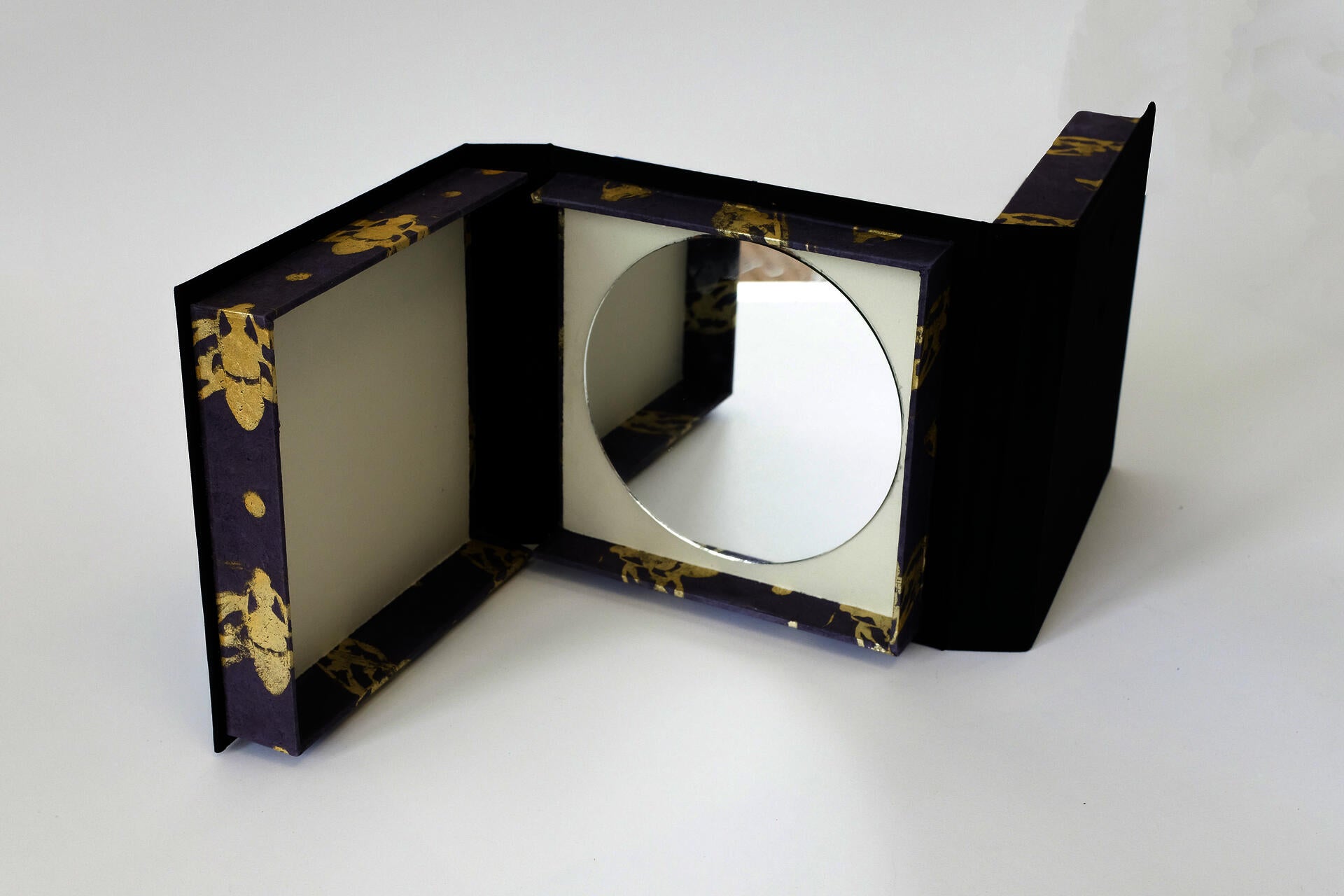Double sided box against a white background that has a mirror inside of it. The edge of the box has a gold moth pattern.