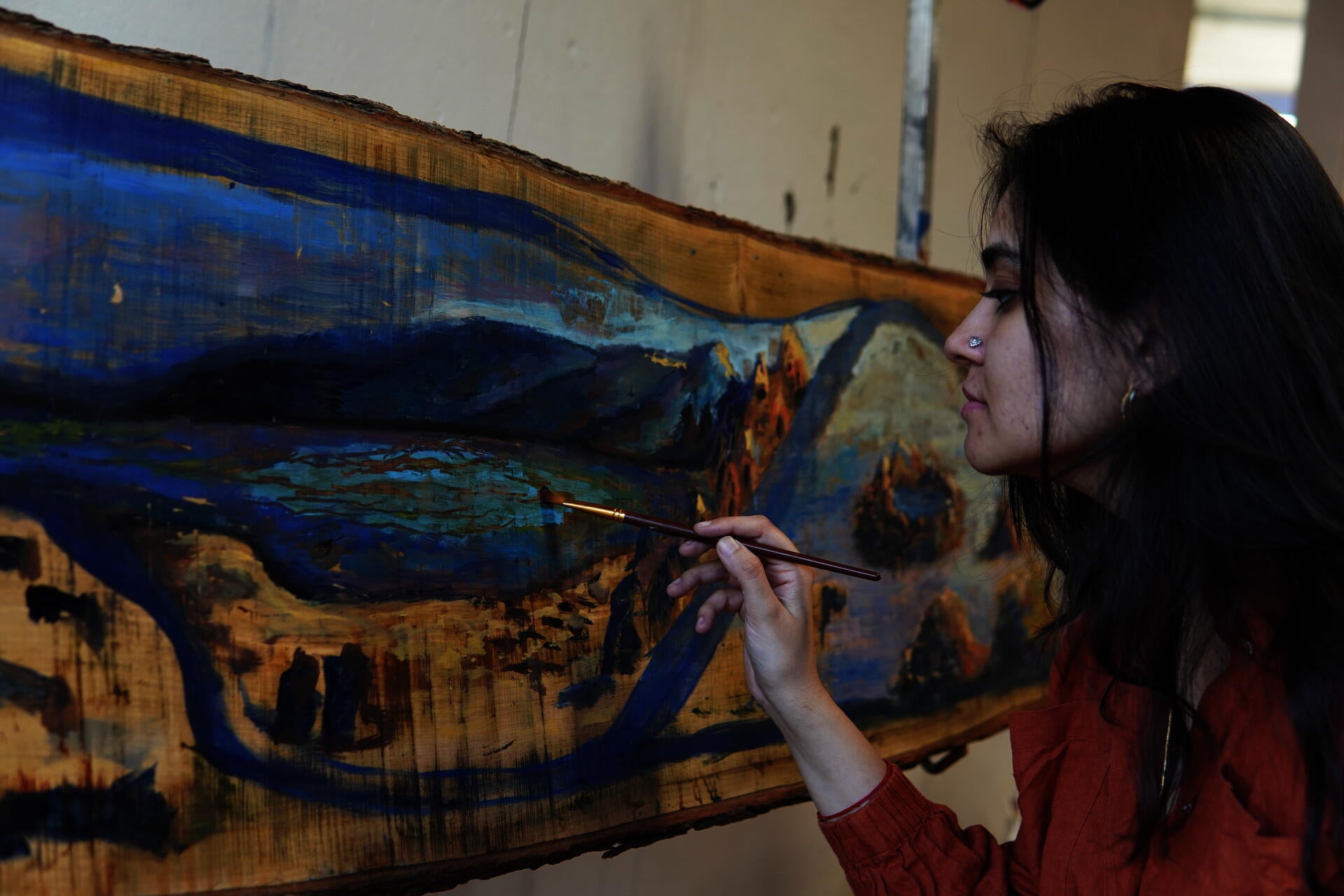 A South Asian woman with long dark hair and a nose ring holds a fine paintbrush close to a large live-edge maple panel propped at an angle. The panel is painted in deep blues, warm ochres, and muted greens