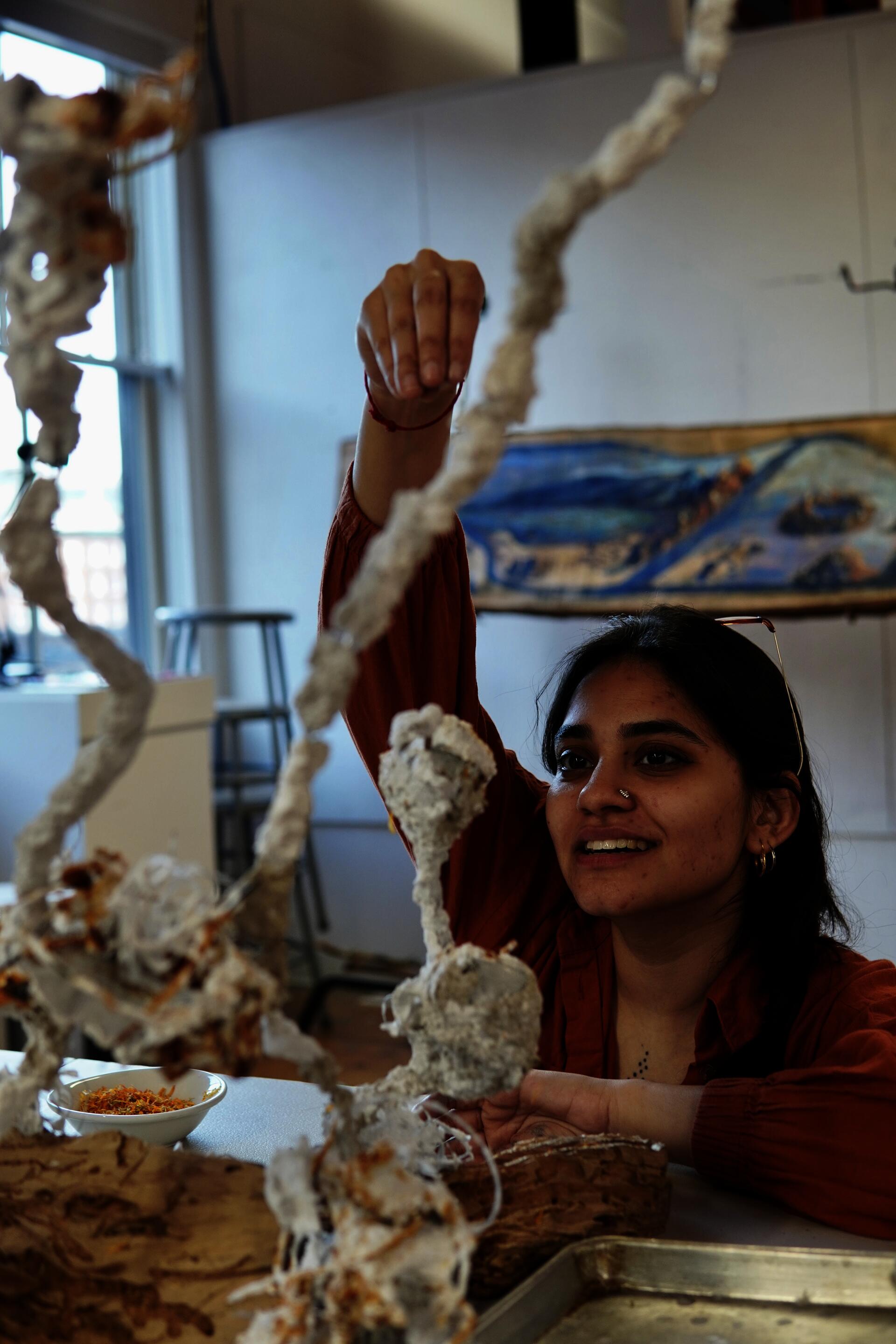 A South Asian woman smiles as she lifts and examines a looping, textured white sculptural form made from paper pulp stuck on a wire armature, rising from a white table surface.