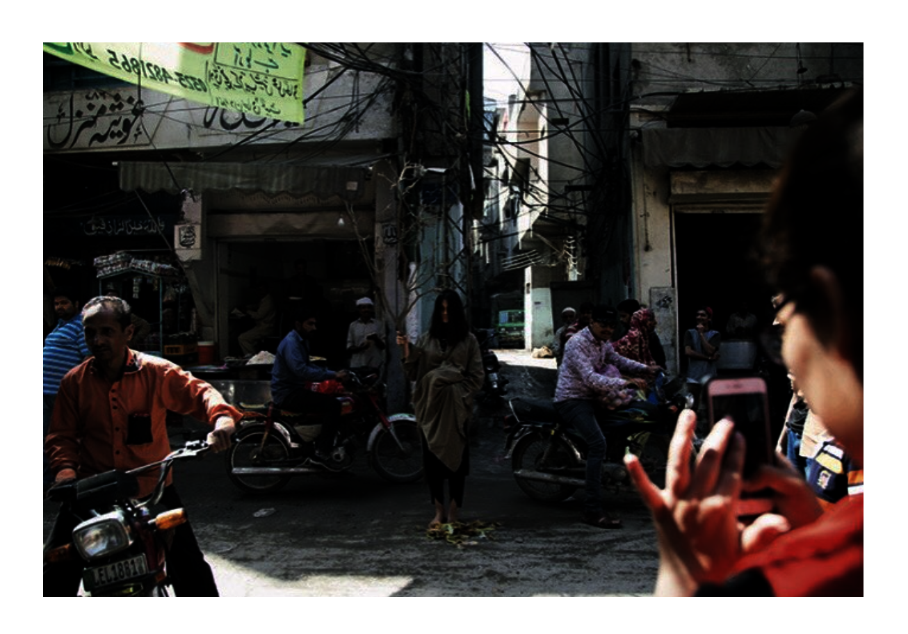 A busy urban street in Pakistan, shot from a slight distance. At the centre of the frame, a figure stands still in a khaki draped garment, barefoot, holding a bare tree upright in the middle of traffic. 
