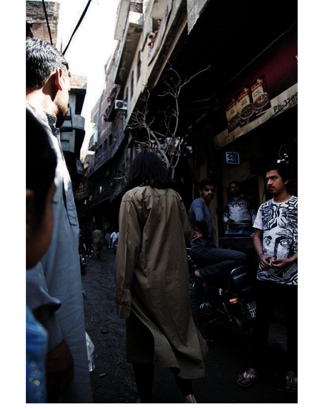 A narrow street in a Pakistani city, photographed from behind. A figure in a long khaki kurta walks barefoot away from the camera into the crowded lane, carrying a bare, branching tree — held upright