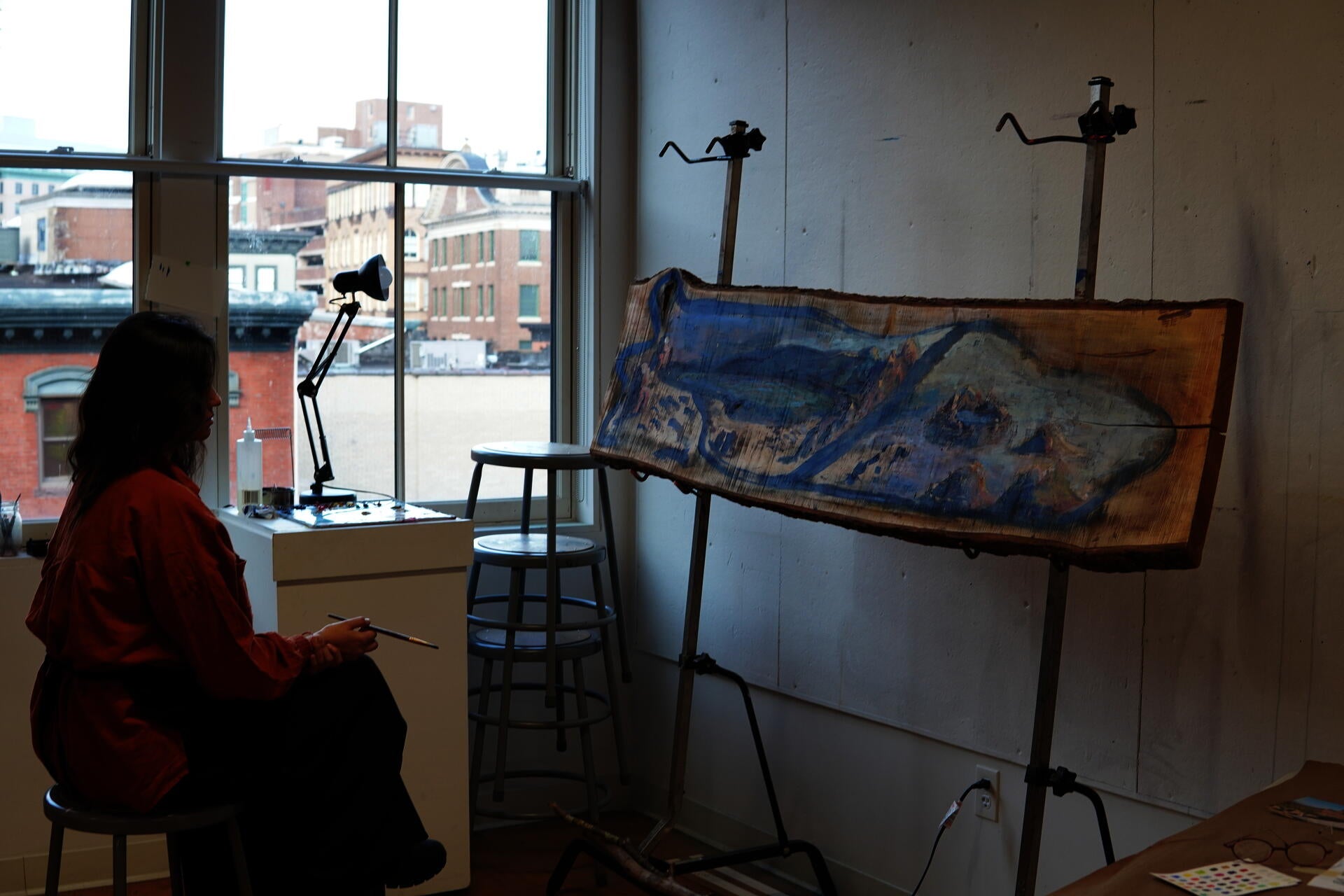 A South Asian woman in a rust-coloured top sits on a metal stool in a high-windowed studio, holding a paintbrush and studying a large live-edge maple panel mounted on a standing easel in front of her.