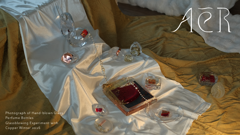 Hand-blown glass perfume bottles arranged on draped white and golden green fabric, each containing small red and copper elements. The composition is softly lit, highlighting transparency, reflections, and material textures.