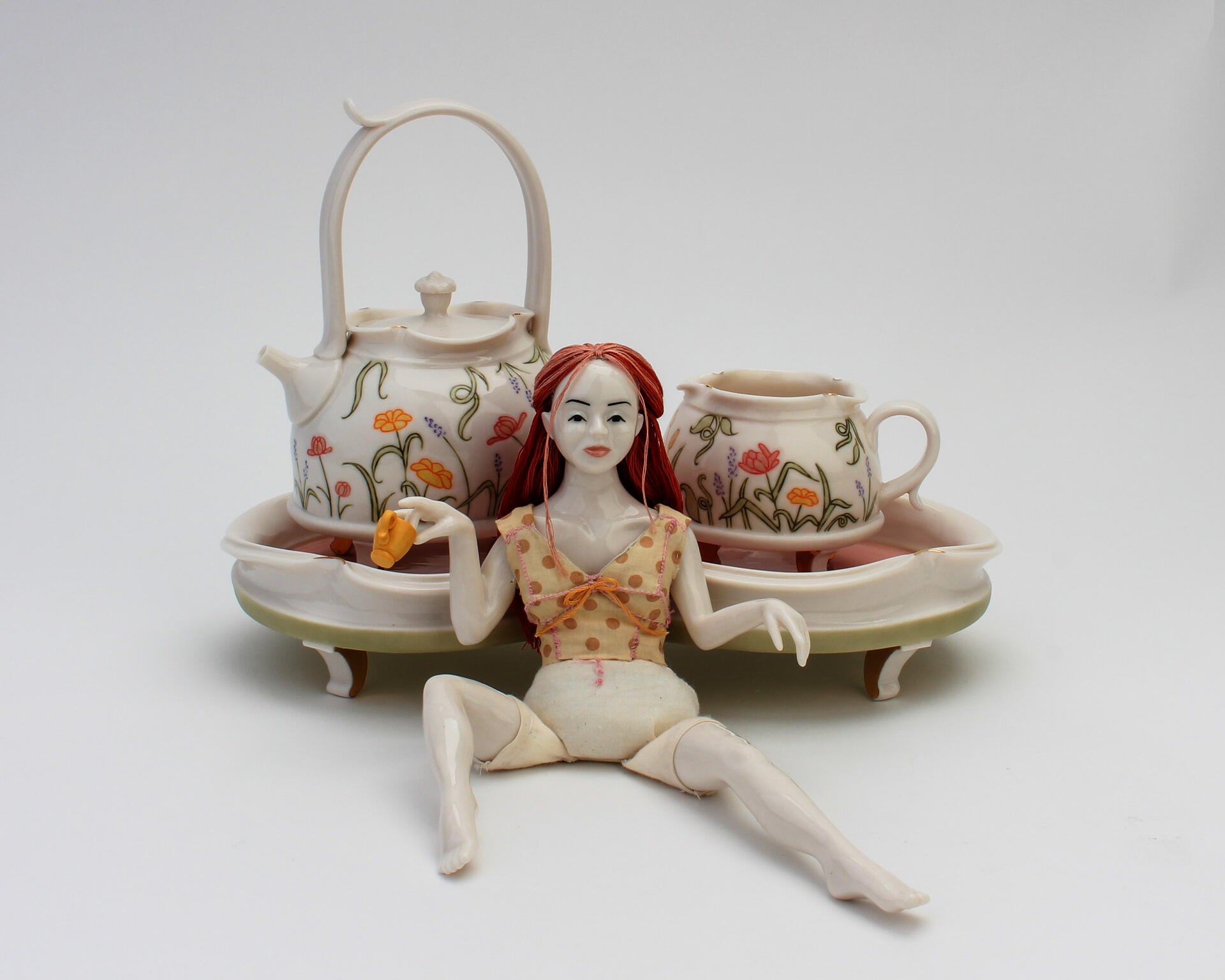 A dainty doll sitting with legs spread apart in front of a human-sized tray, teapot, and teacup decorated with floral designs
