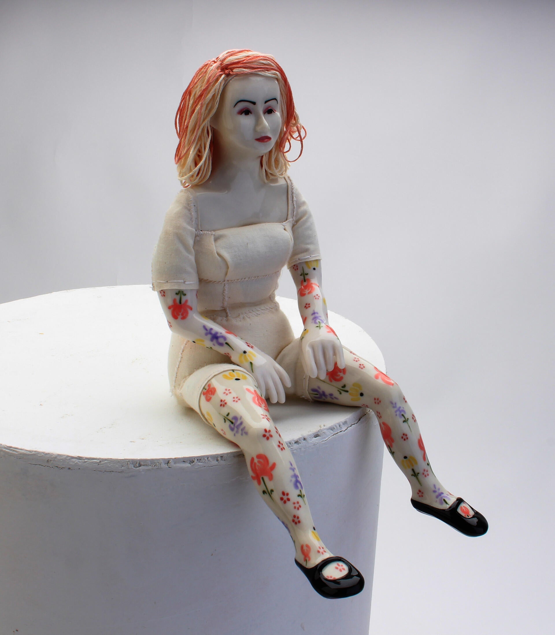 A porcelain doll with white clothes, pink hair, and floral designs on arms and legs sitting on the edge of a pedestal