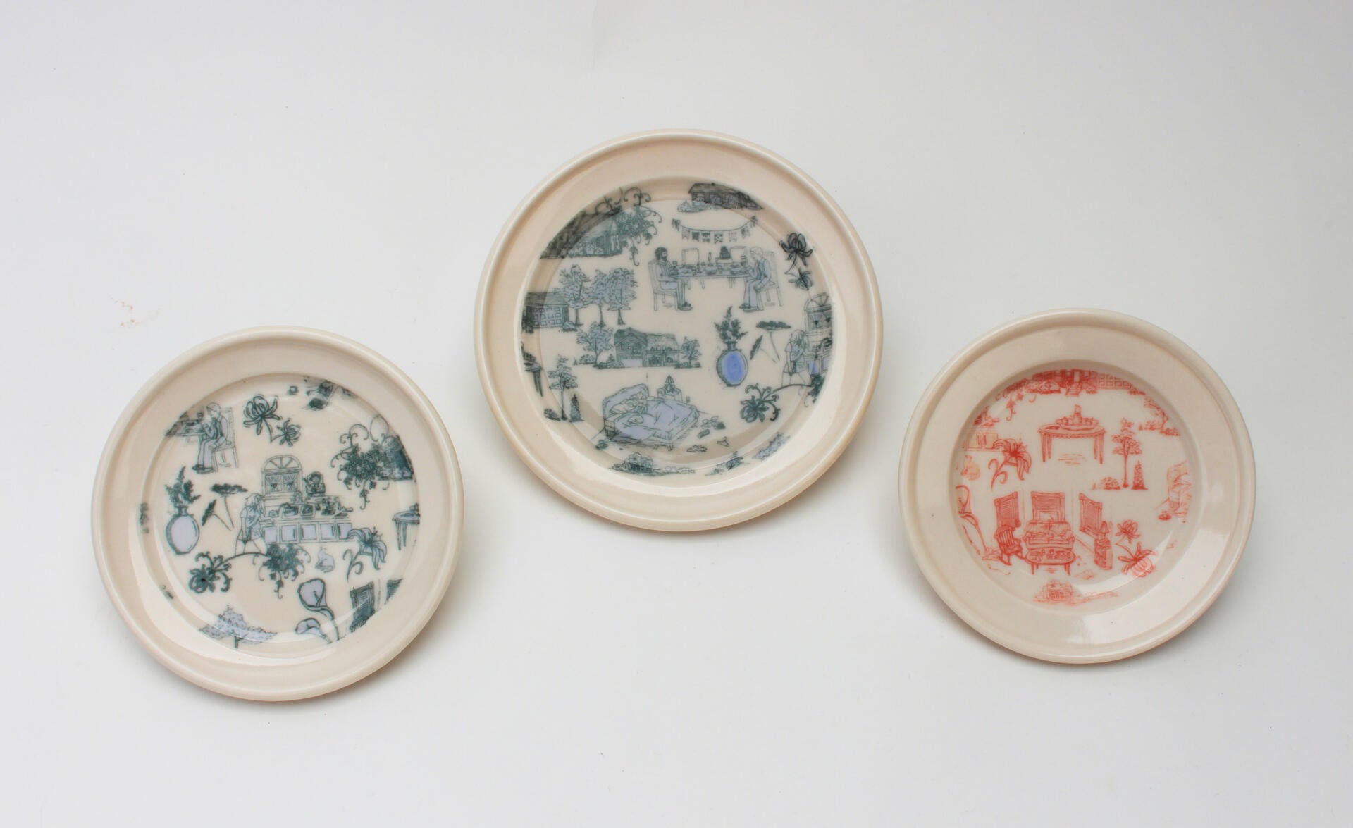 Two white plates with a blue pattern of illustrated cluttered domestic scenes and one white plate with the same pattern in pink