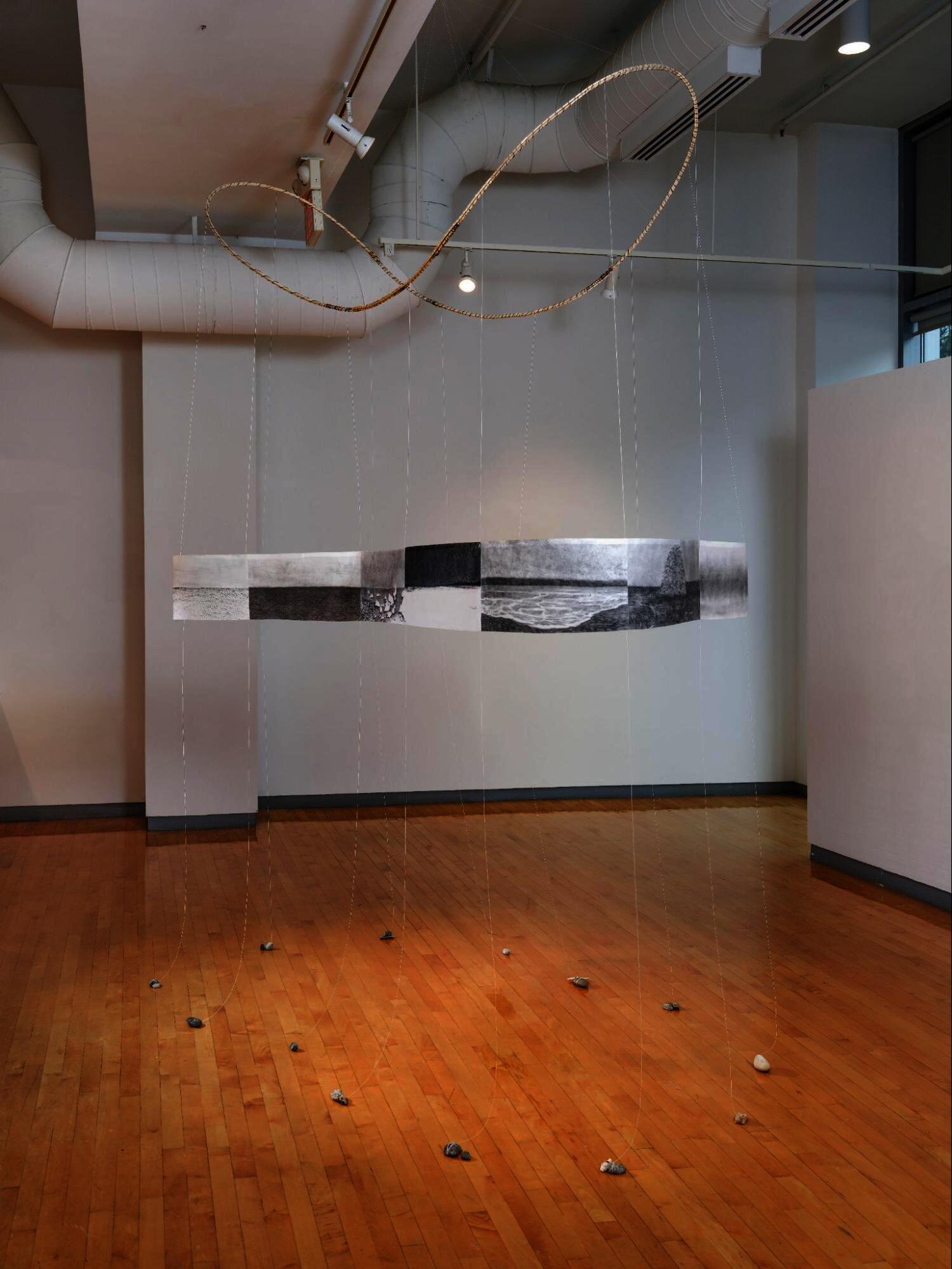 Suspended circular band of drawings hung by thin strings, with stones on the floor beneath in a gallery space