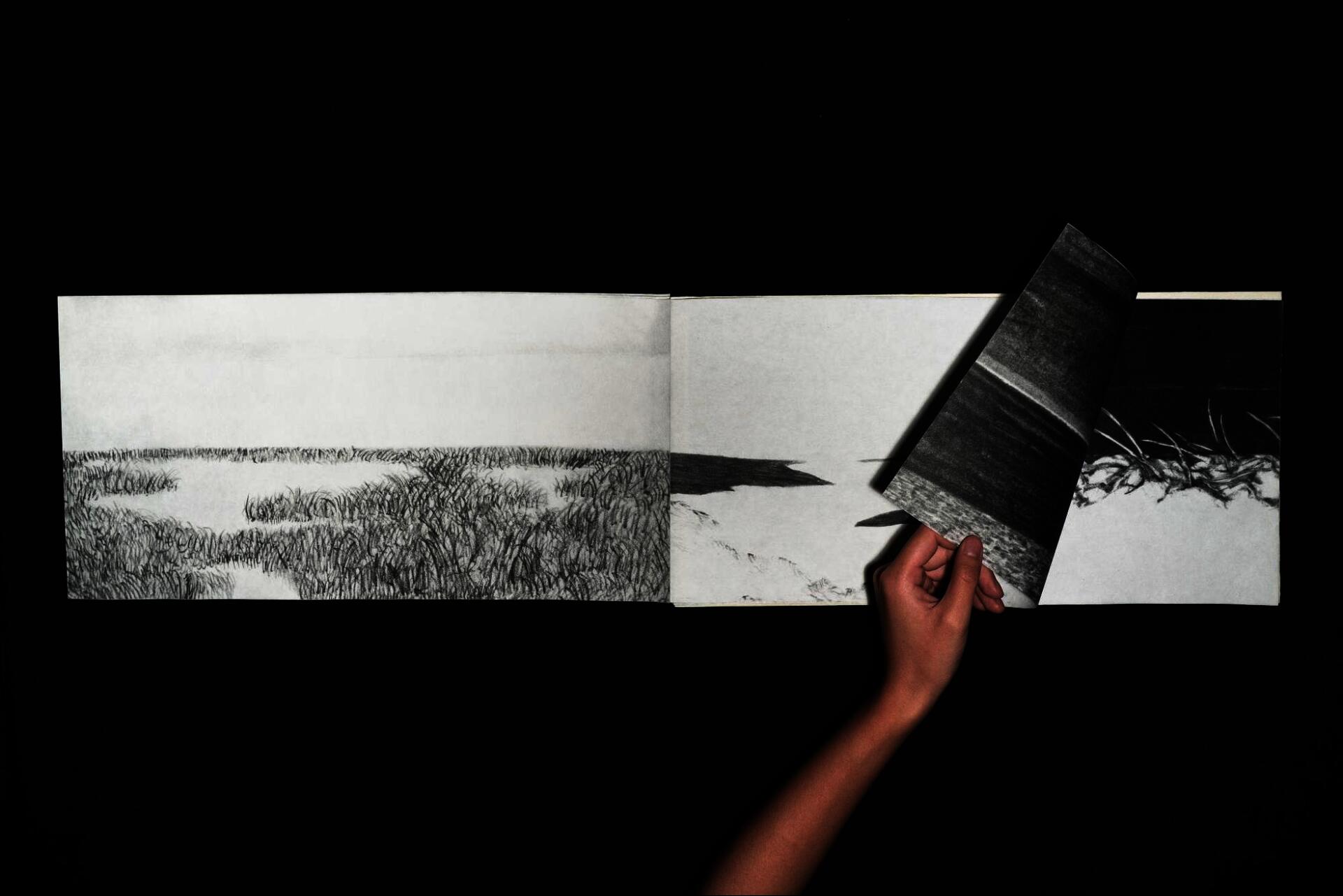 Hand lifting a page of an accordion book revealing a continuous charcoal landscape drawing across connected panels