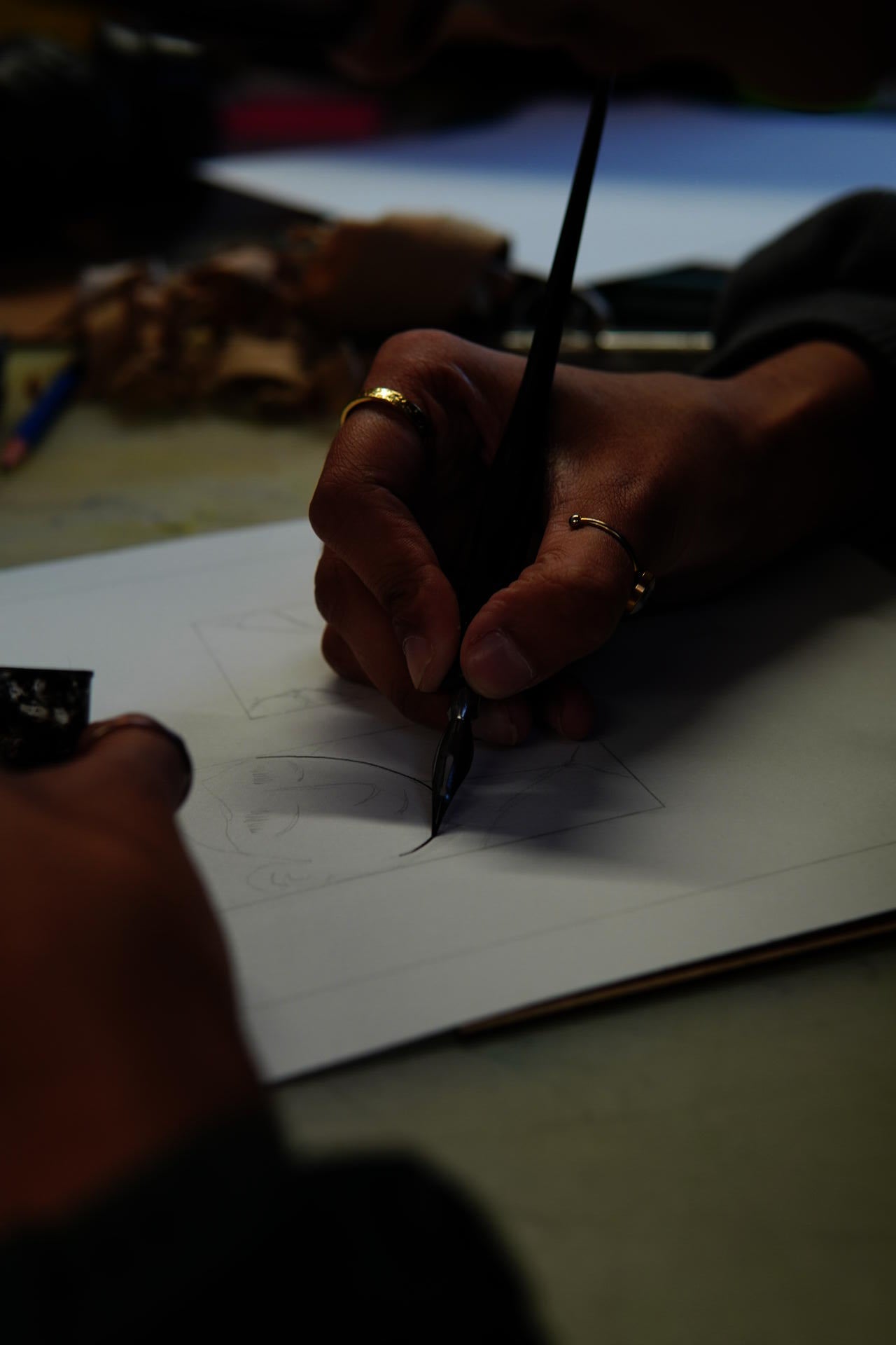 Image shows artist inking a hand-drawn comic page on paper, applying ink over pencil lines with a dip pen as part of the final linework stage of comic production.