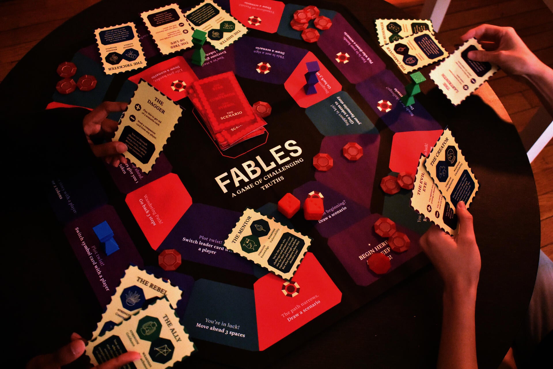 A colorful board game layout displays "Fables: A Game of Challenging Truths" with character archetype cards, scenario cards in red and purple, coral-colored tokens, and red dice, arranged on a wooden table with players' hands holding cards.