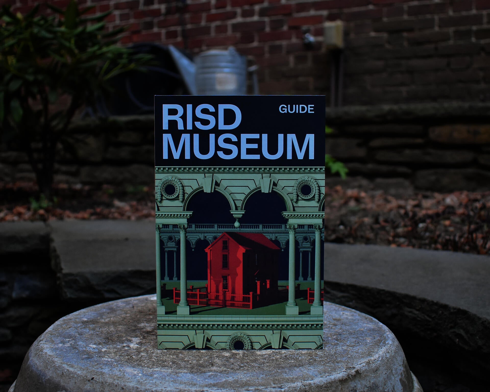 A RISD Museum Guide cover stands upright on a stone surface, featuring a navy blue header with light blue typography and an illustration of the museum's red brick architecture framed by ornate green classical detailing.