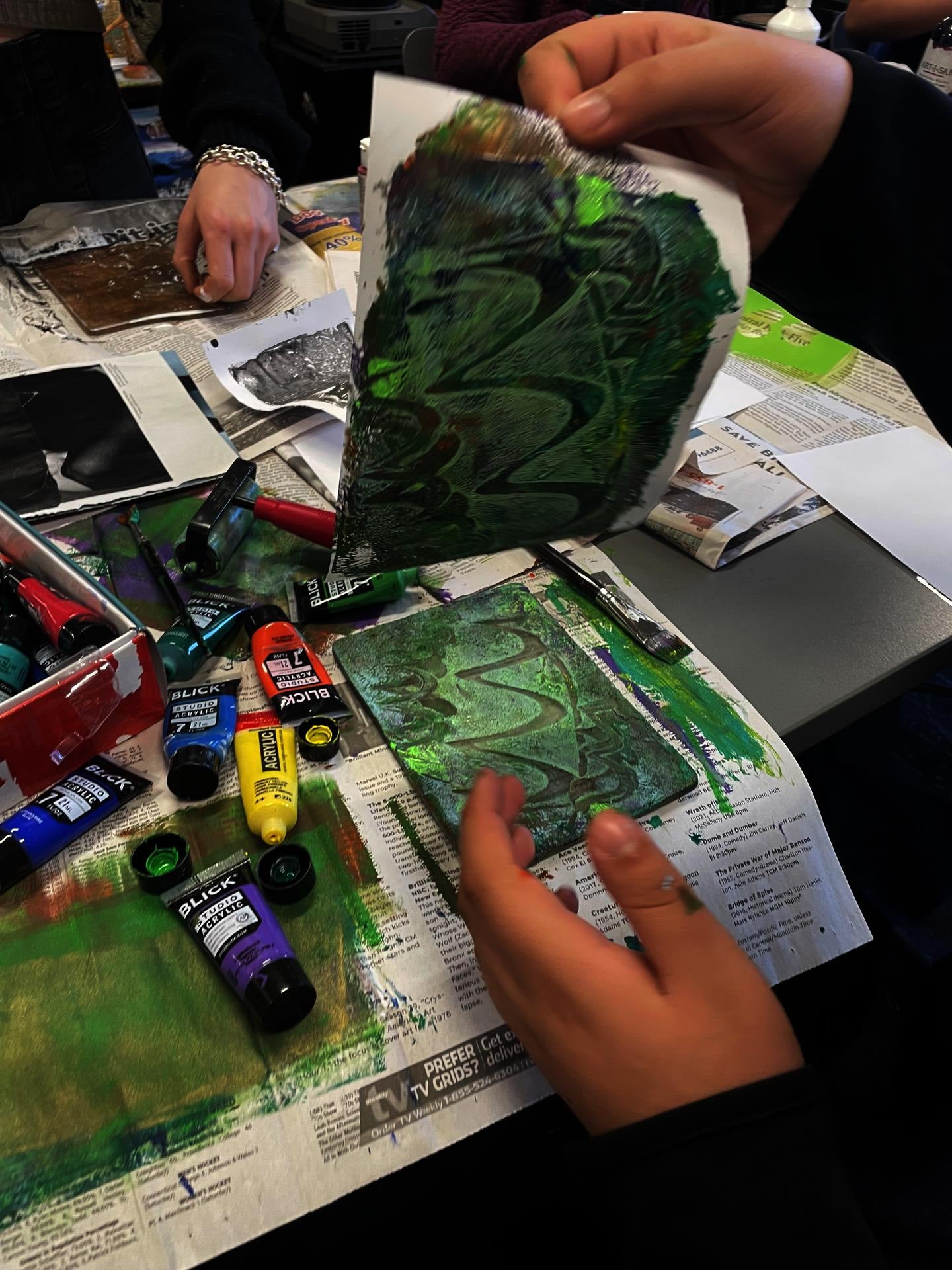 student lifting green abstract image on paper from a geli plate on top of a table covered in paint tubes and newspaper.