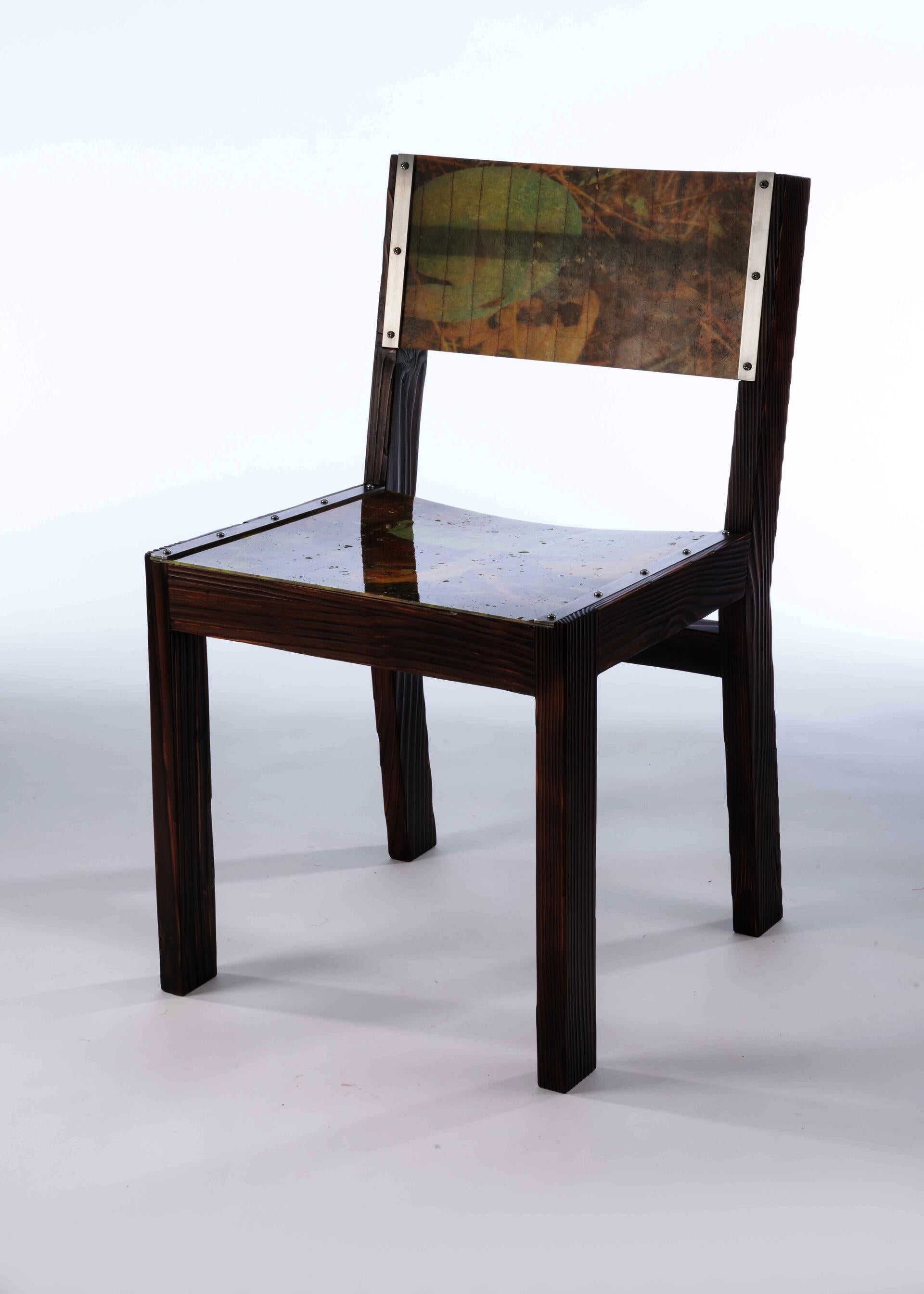 A chair with a blocky frame made of darkened burnt wood with a seat and back panel made from fiberglass that has images of the forest floor laminated inside.