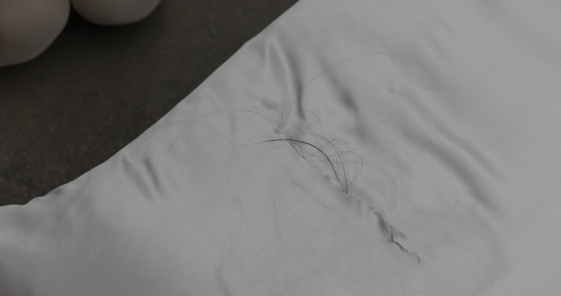 Close view of a white pillow on concrete floor with a small tear revealing strands of dark hair emerging from inside; smooth fabric contrasts with rough ground.