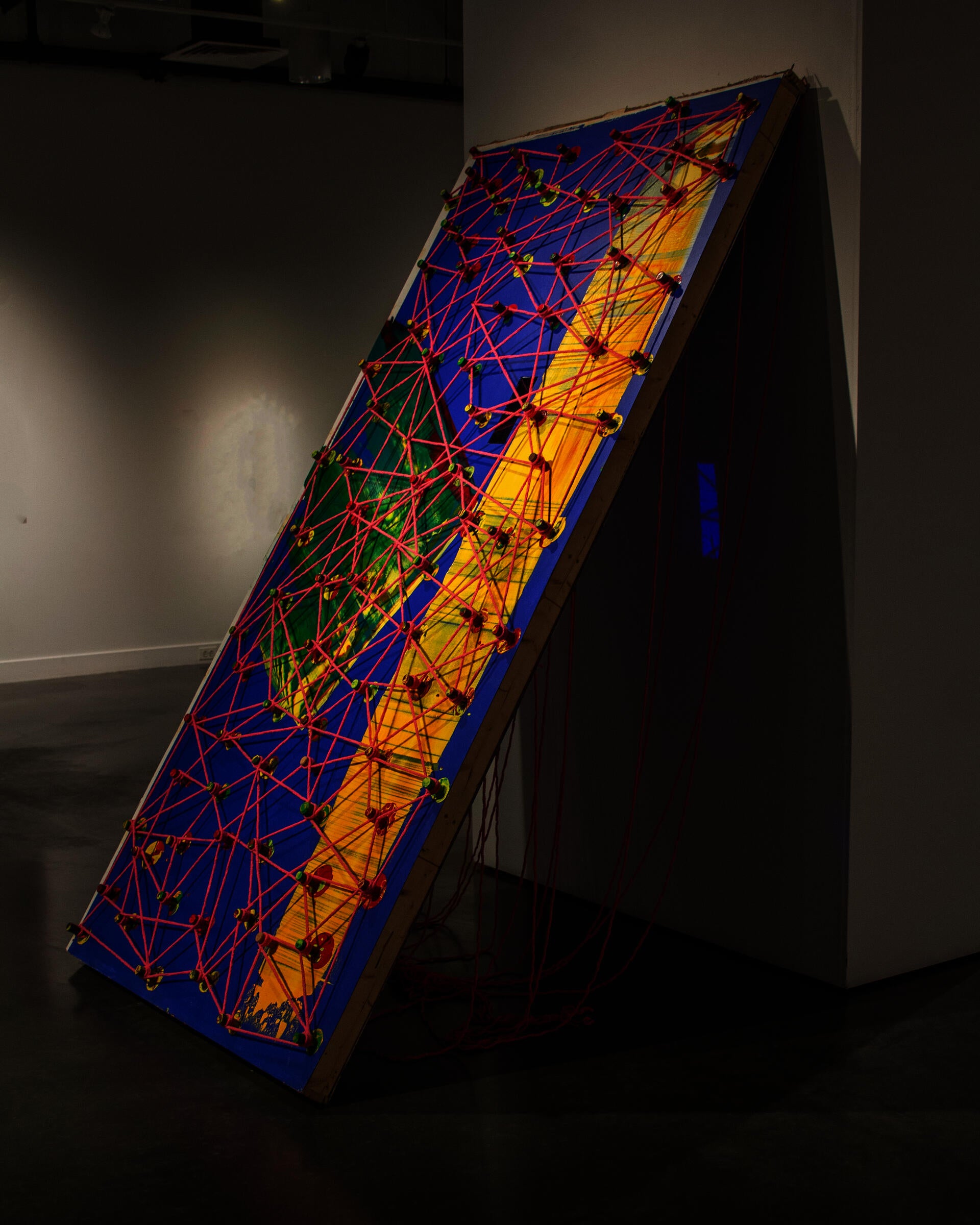 This represents a big blue wooden panel, stand diagonally against the wall. It has long yellow streak, with wine many corks. all the wine corks are connected through a pink yarn, represents an abstracted language.