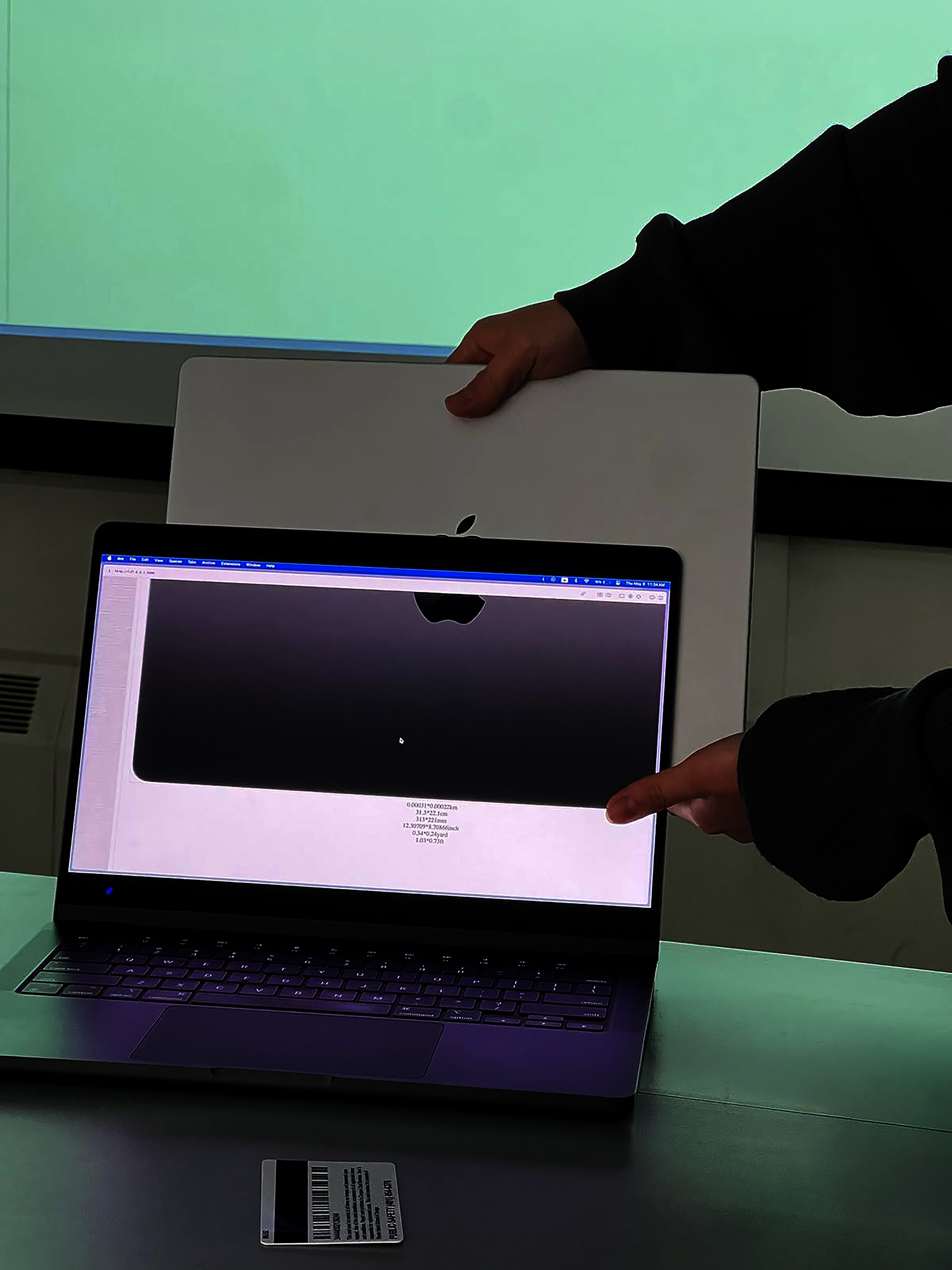 Two hands hold a closed silver laptop behind an open laptop displaying a grayscale scan of its back. A barcode card rests on the table in front of the screens.