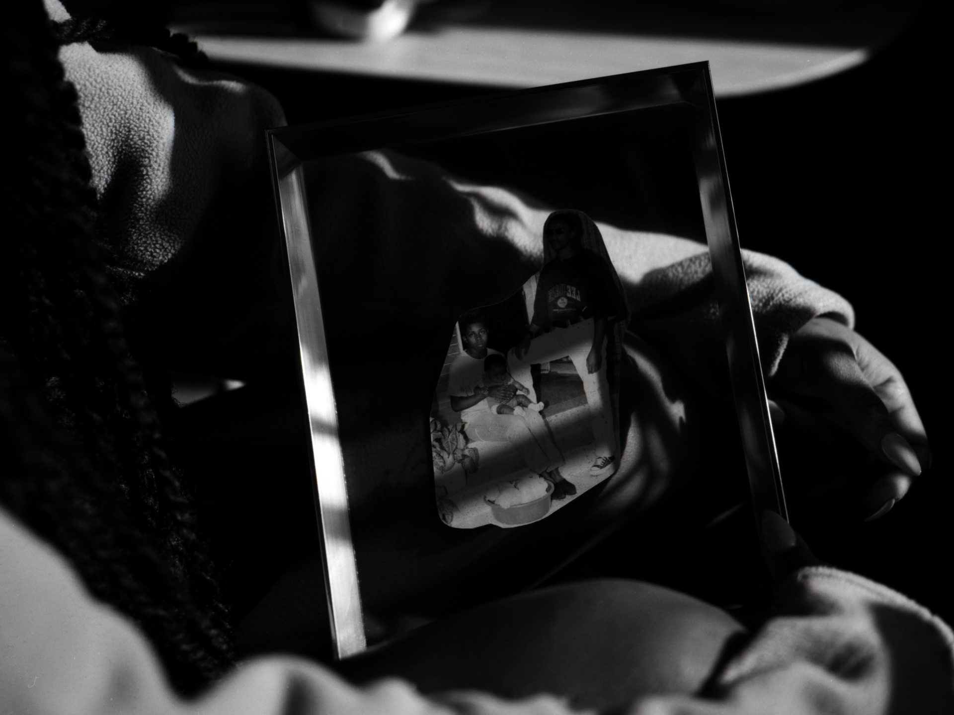 Black and white photograph of a person holding a framed photograph with a torn collage of people in casual attire. There are three figures. Sunlight casts shadows across frame.