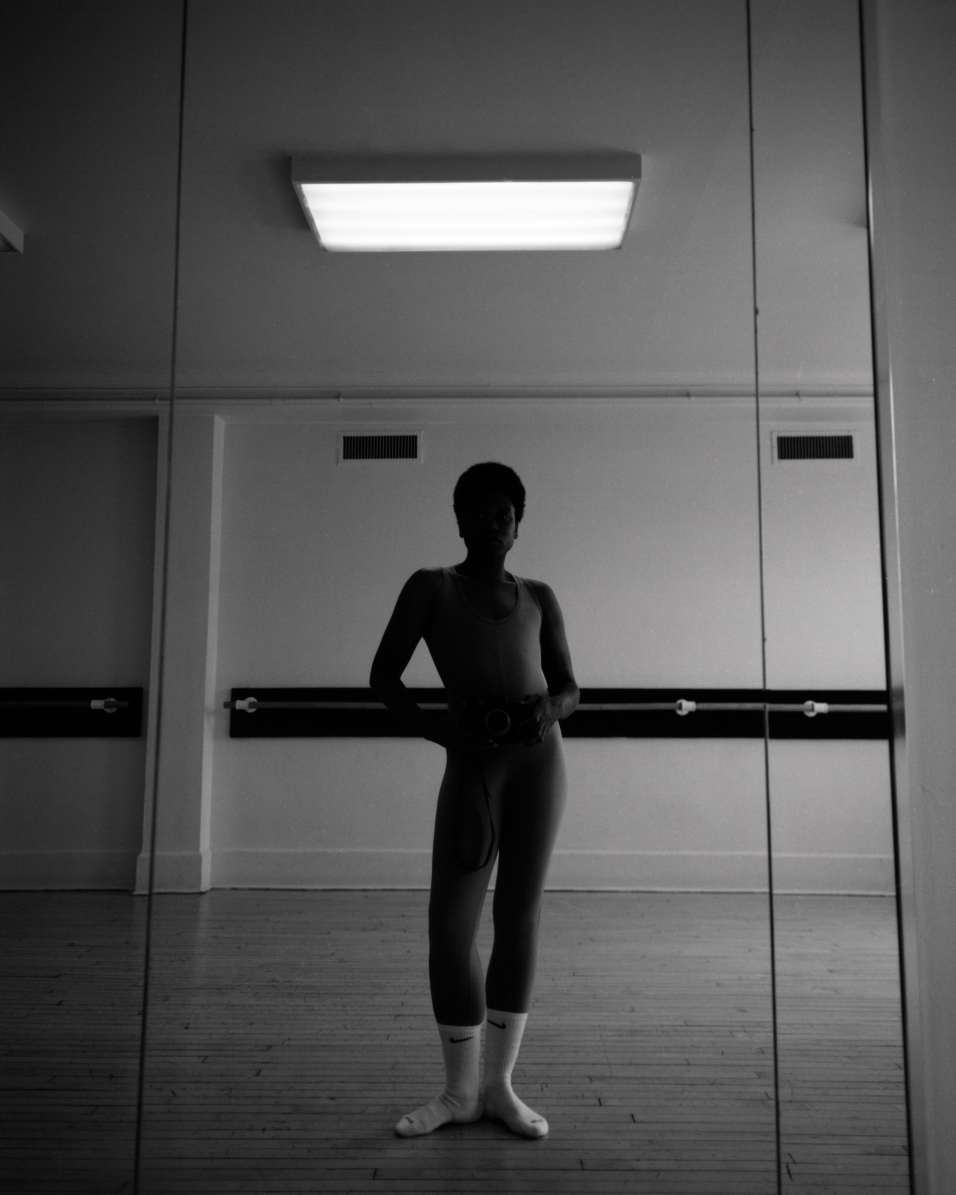 Black and white photograph of a person in a leotard and socks, holding a camera in a mirrored dance studio. The space is minimal and reflective.