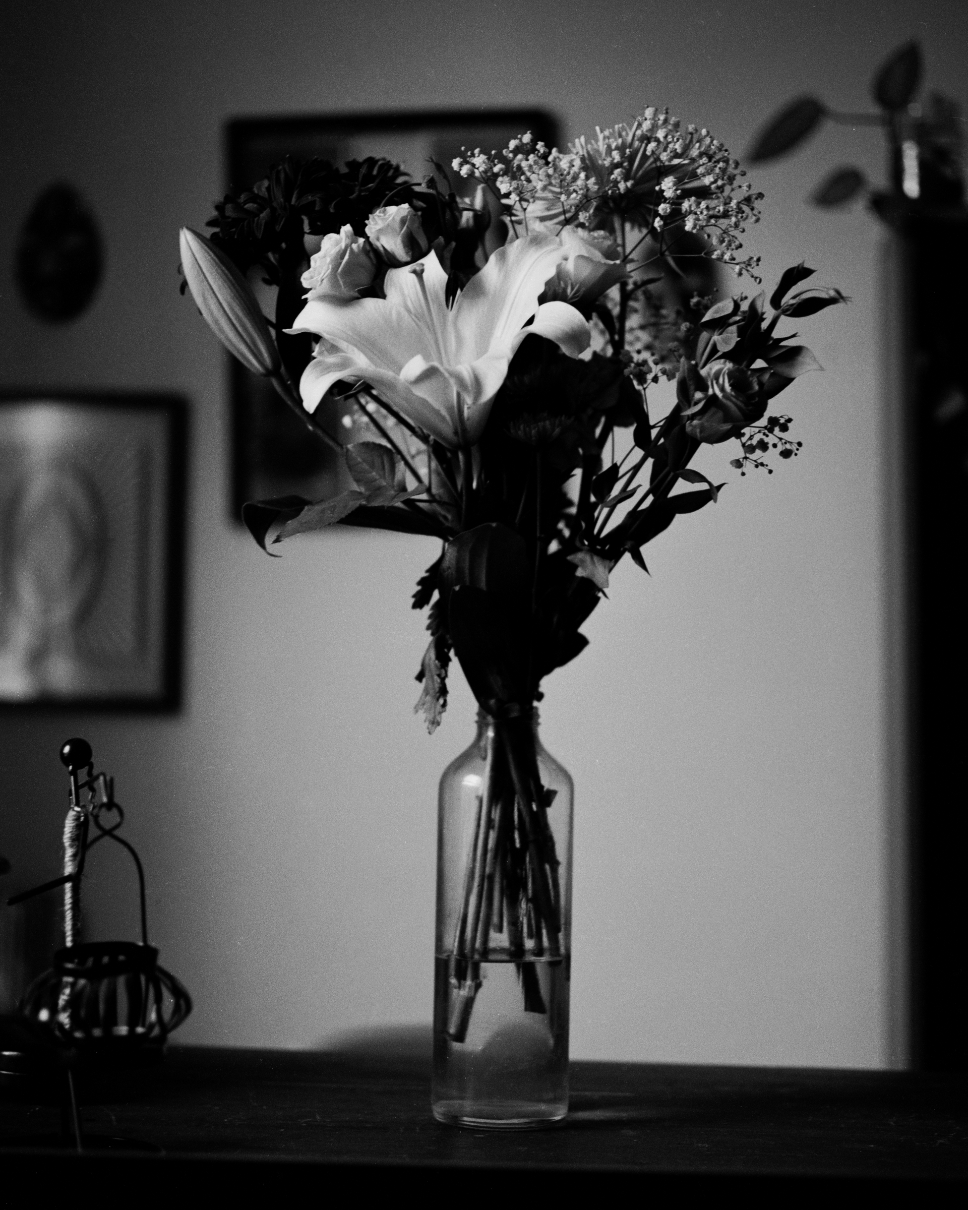 Black and white photograph of a bouquet of lilies and roses in a tall glass vase on a wooden table.