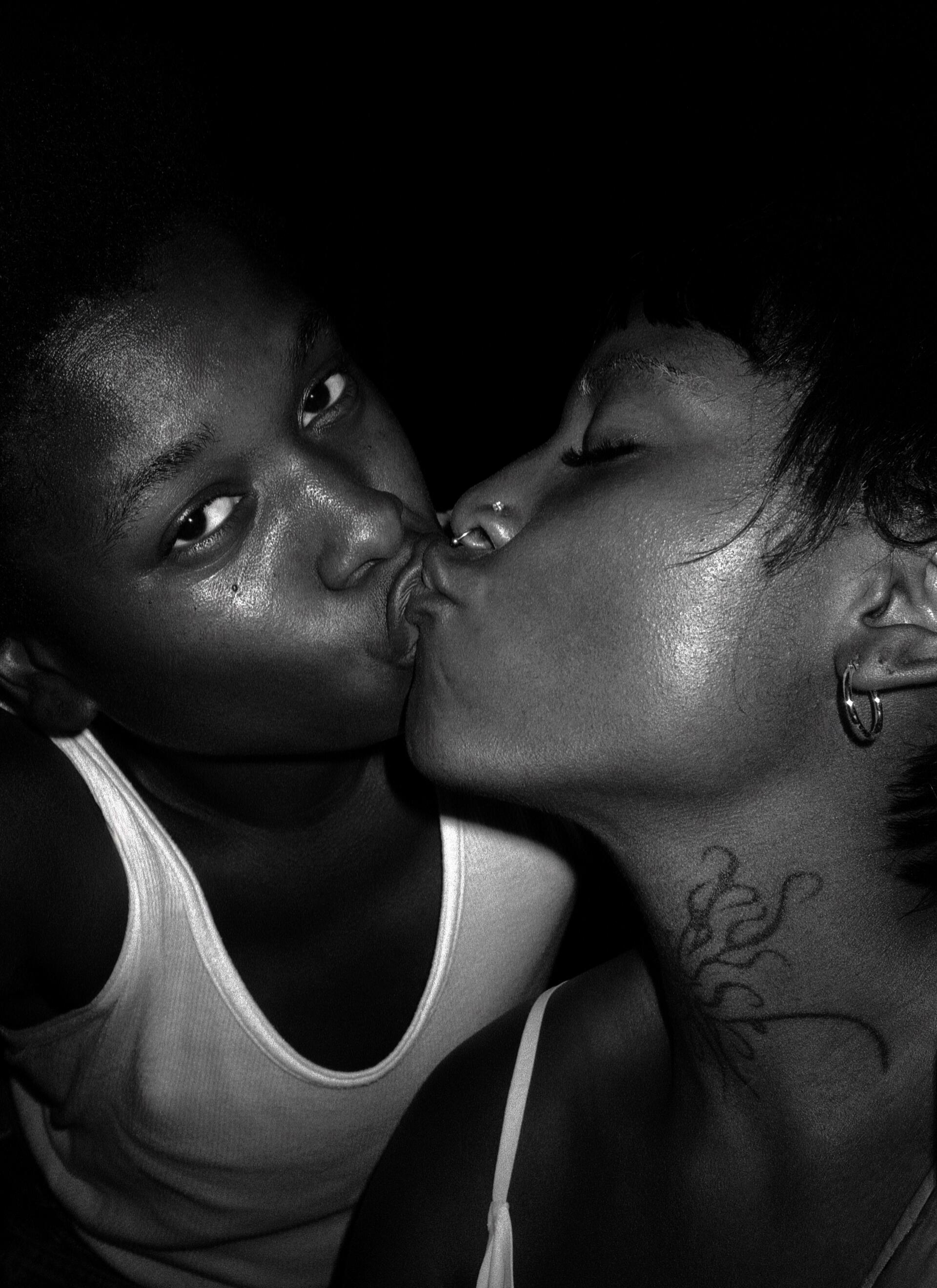 Black and white photograph of two people kiss intimately, their faces close. Person on the left is looking directly into the camera and person on the right has a tattoo on their neck, wearing hoop earrings.