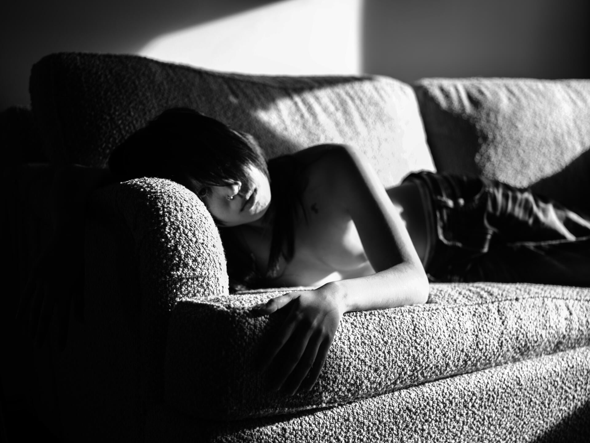 Black and white photograph of a person with long hair lounging on a textured sofa in soft sunlight. Their eyes are almost fully covered from their bangs.