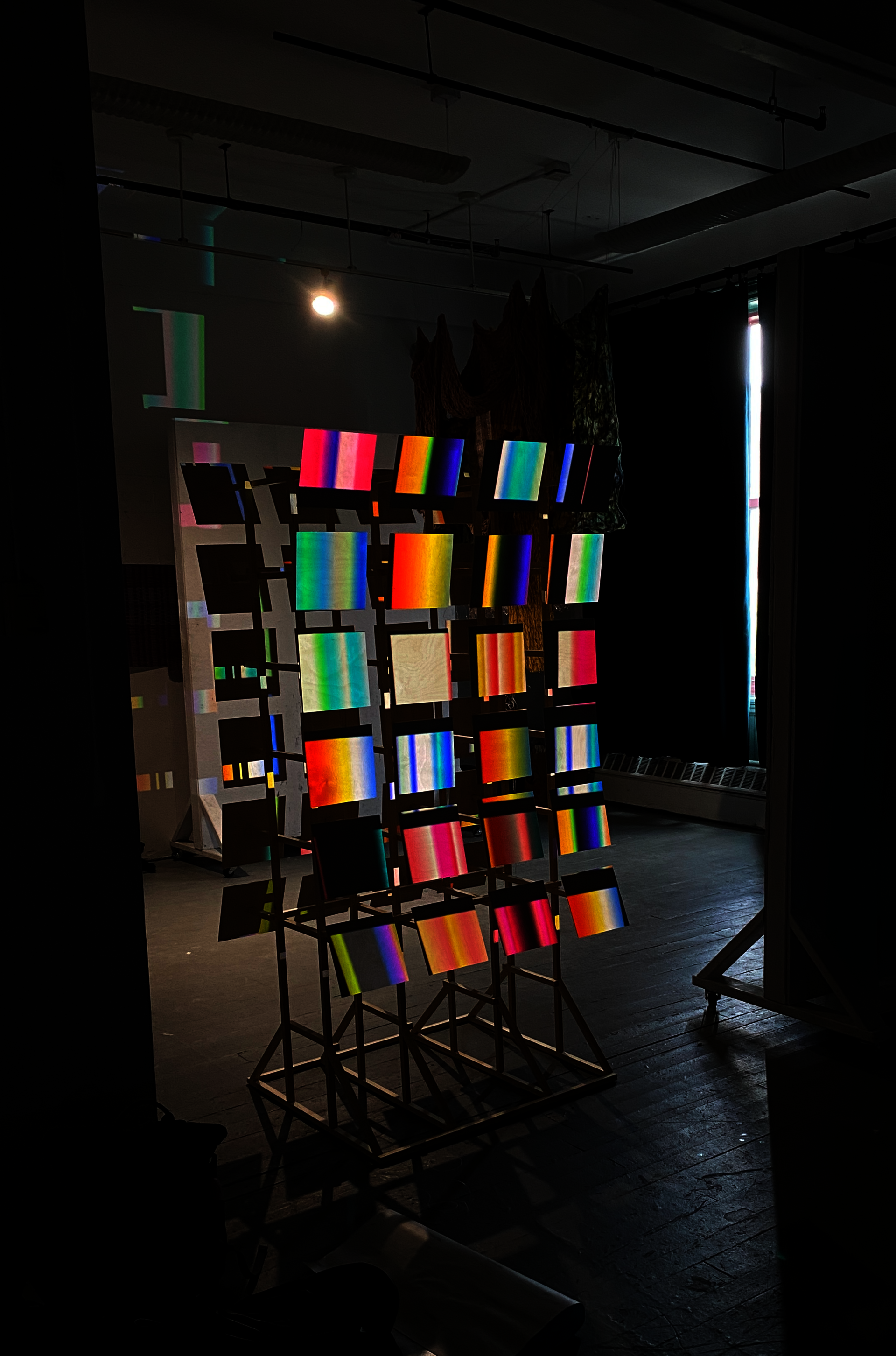 Dim installation view of a wooden structure holding many glowing gradient panels, with projected color bands and shadows filling a studio space.