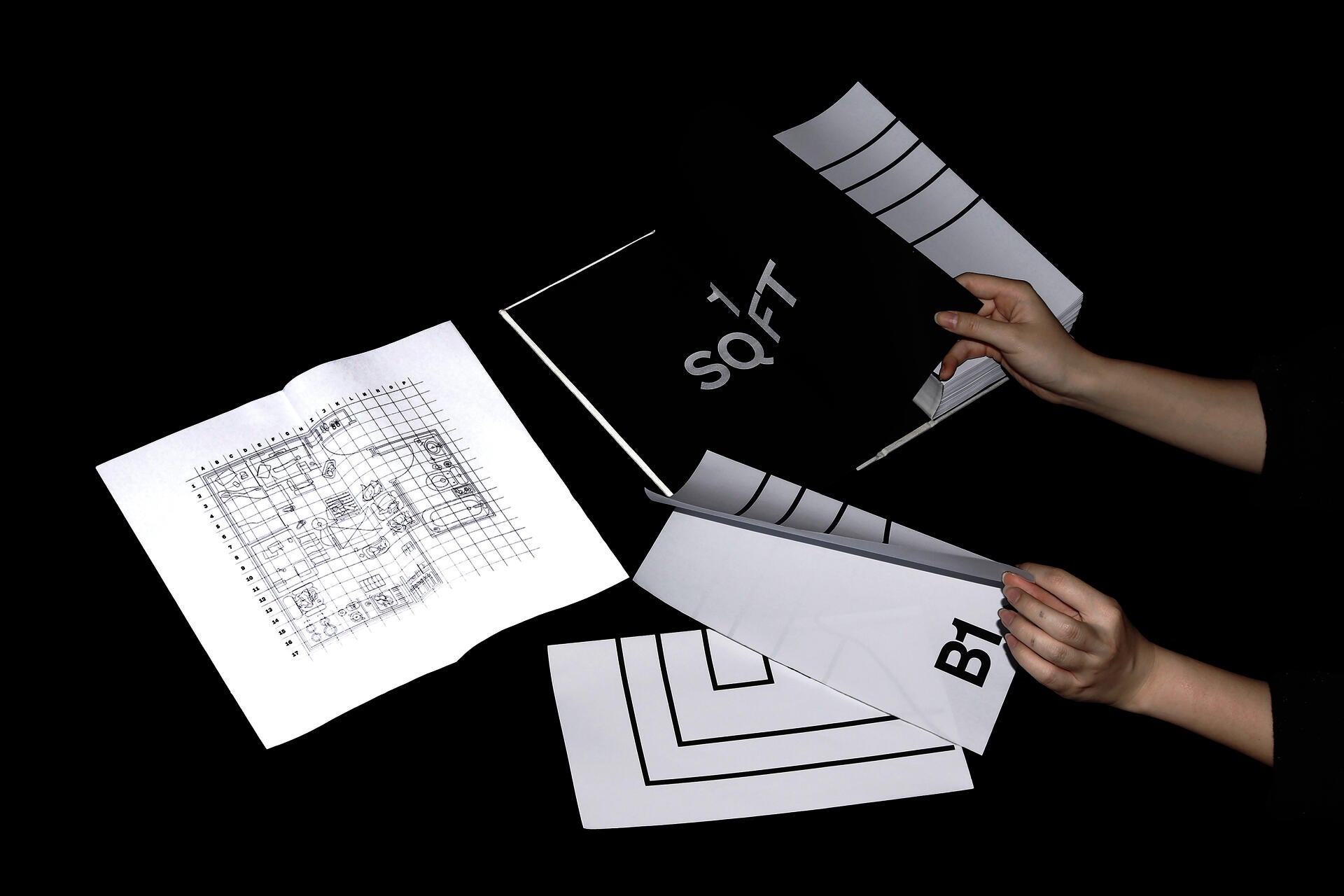 Hands unfold a large black-and-white book-object, revealing spreads with floor-plan grids, thick contour lines, and large room labels against a black studio background.